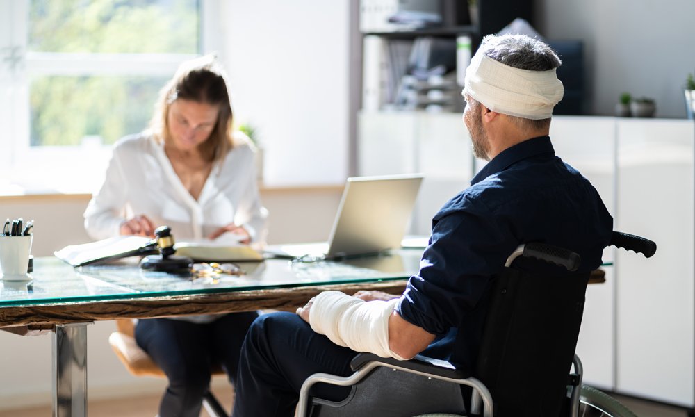 A man with a bandaged head and arm, in a wheelchair, is shown from behind facing a woman who is seated at a desk with a laptop and legal documents.