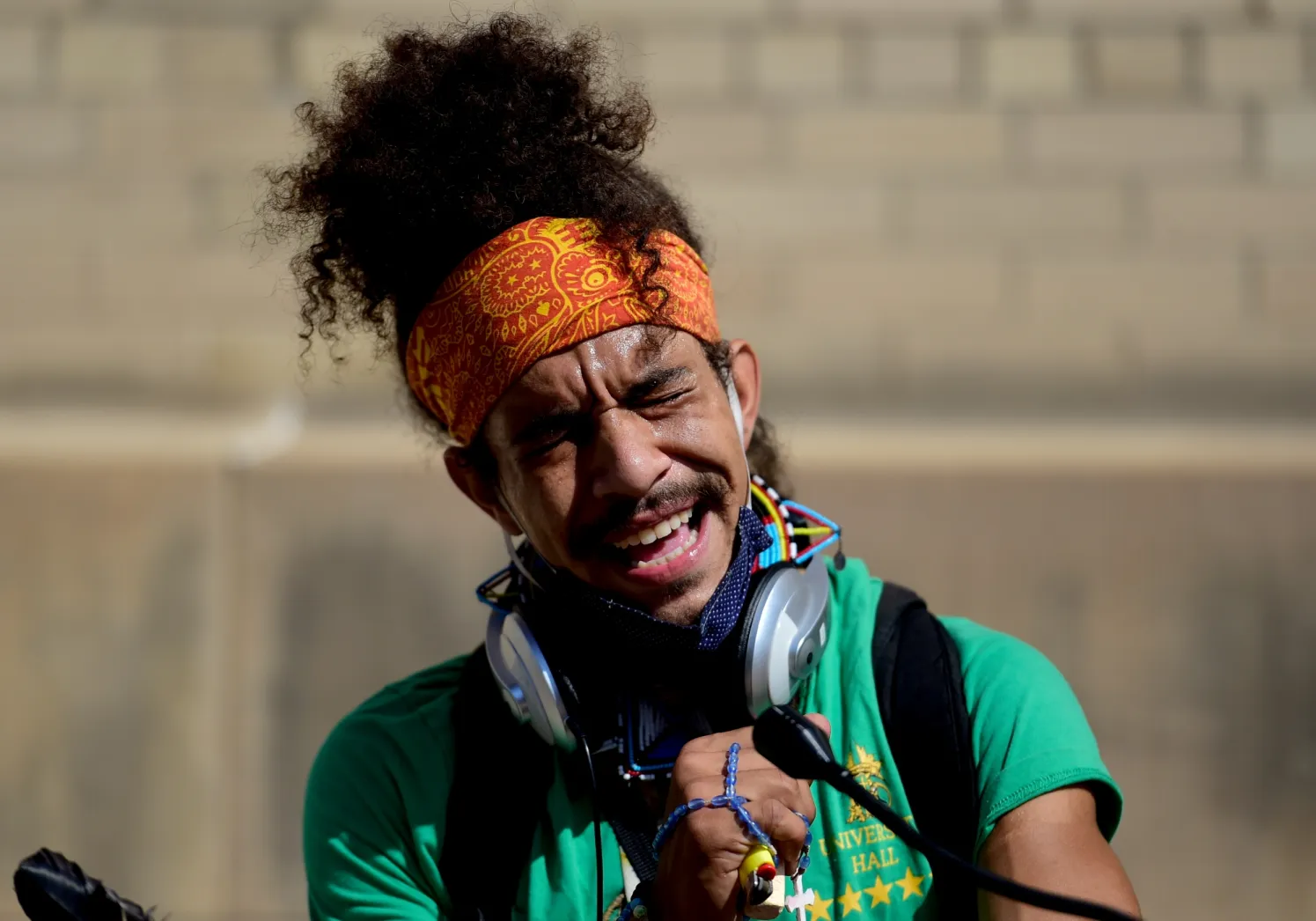 A person with curly dark hair and an orange patterned headband, wearing a green shirt and headphones around their neck, is speaking into a microphone with their eyes closed and mouth open in an expression of emotion or speech.