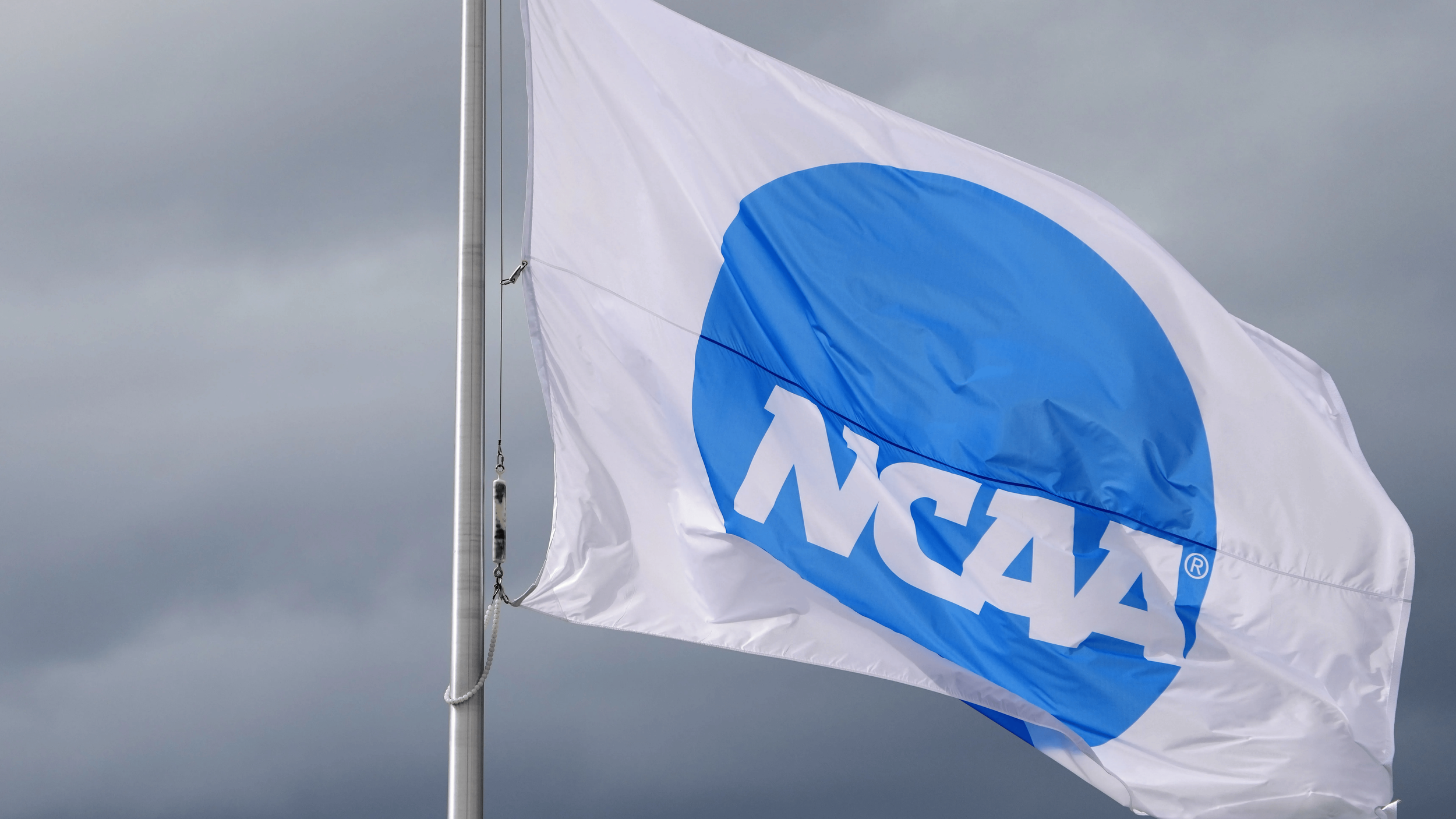A white flag with a blue circle and "NCAA" logo in white letters flies against a cloudy grey sky.