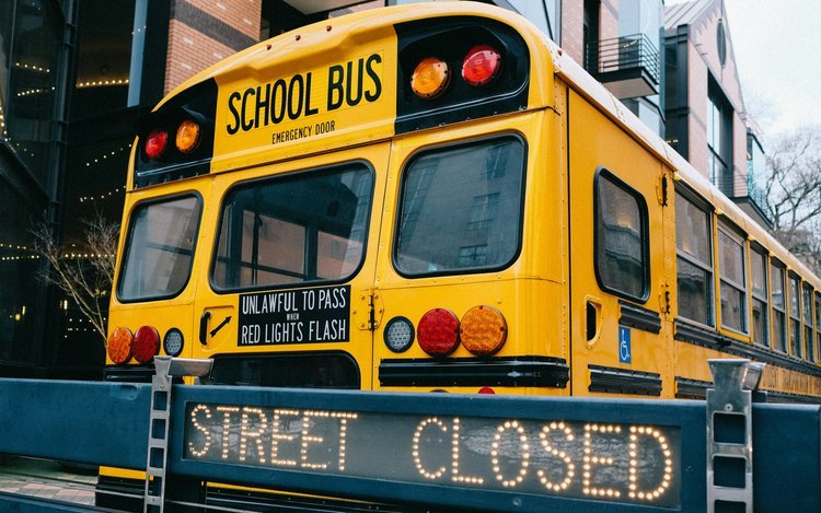 A bright yellow school bus is parked behind a "STREET CLOSED" digital sign, with buildings visible in the background.