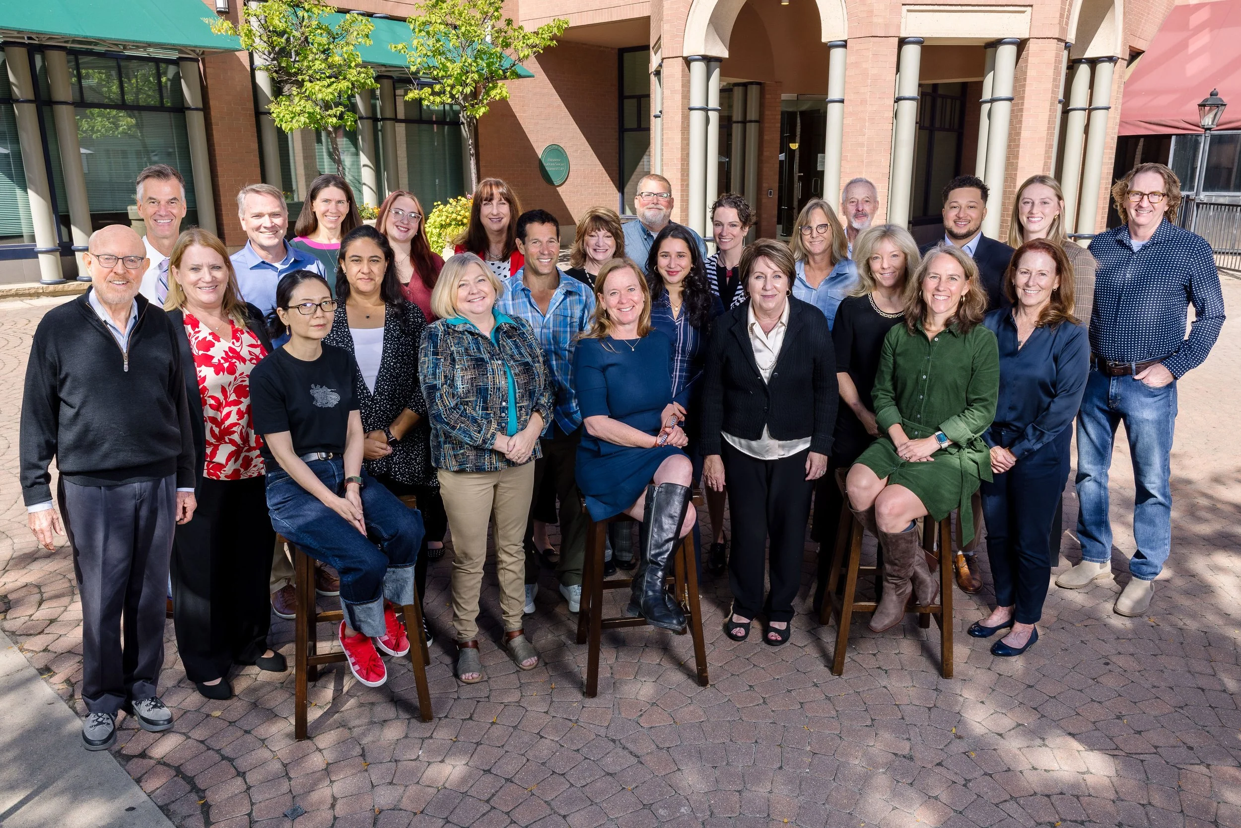 A group photo of a diverse staff of approximately thirty people, some seated on stools and others standing, smiling outdoors in an architectural plaza with brick pavement and archways in the background.