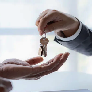 A hand exchanges a set of keys with a house-shaped keychain to another hand, while a contract and a miniature house model are visible on the table, symbolizing real estate transactions.