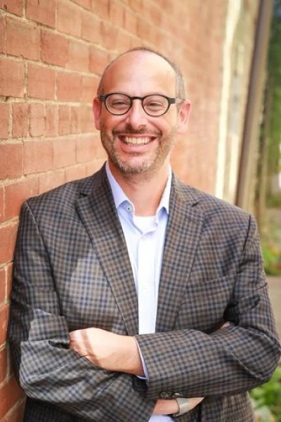 professional headshot of man with glasses leaning against brick wall