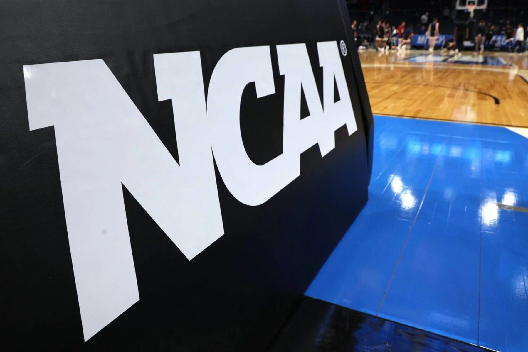 A close-up of a black and white NCAA logo on a padded court-side barrier, with a basketball court visible in the blurred background.