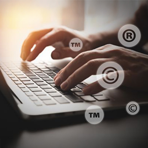 A person's hands are typing on a laptop keyboard, with various intellectual property symbols (TM, R, C) overlaid as translucent icons, suggesting digital work related to trademarks or copyrights.