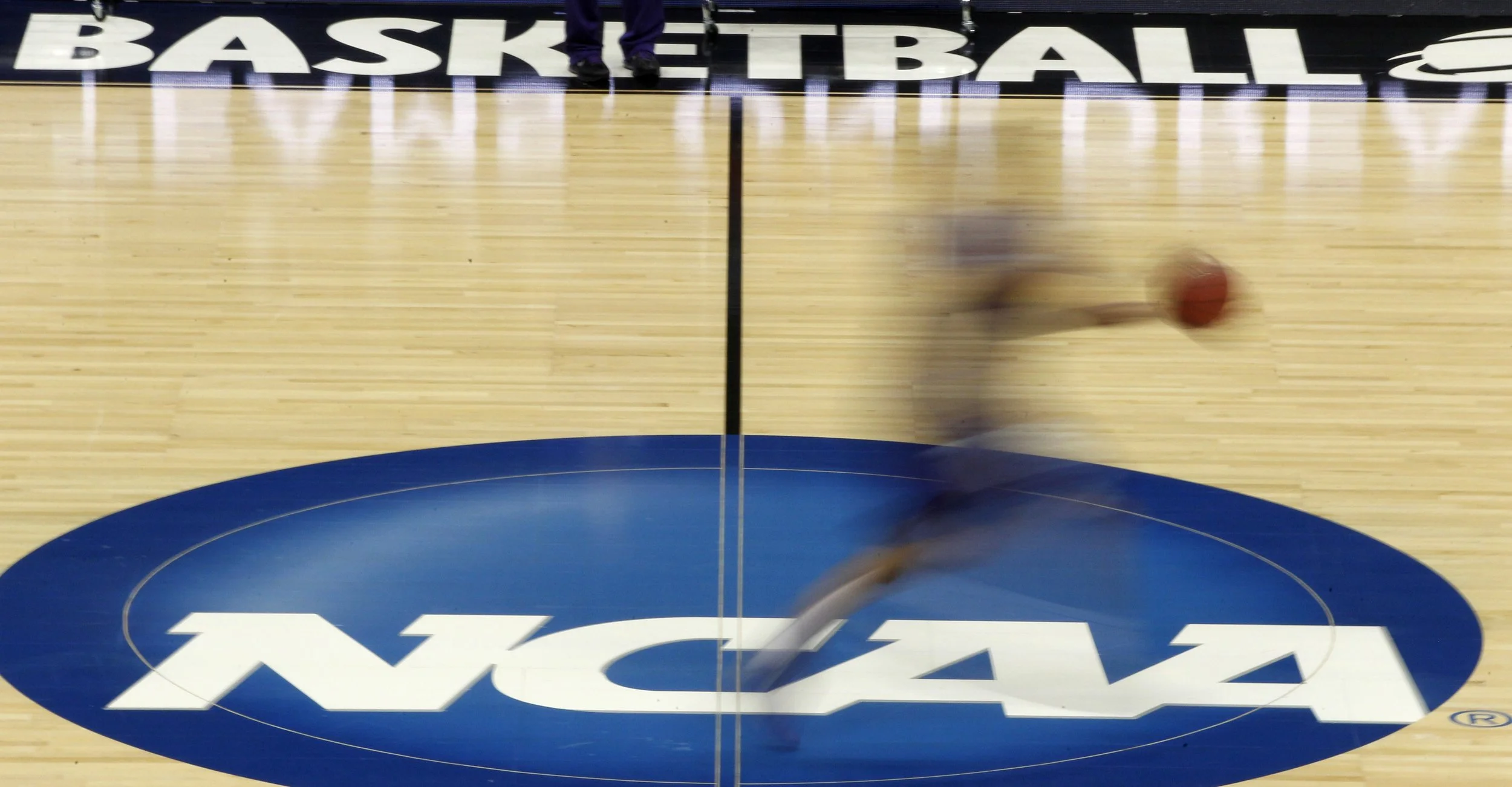 An overhead view of a basketball court with the NCAA logo at center court and "BASKETBALL" text on the sideline, showing a blurred player in motion with a basketball.