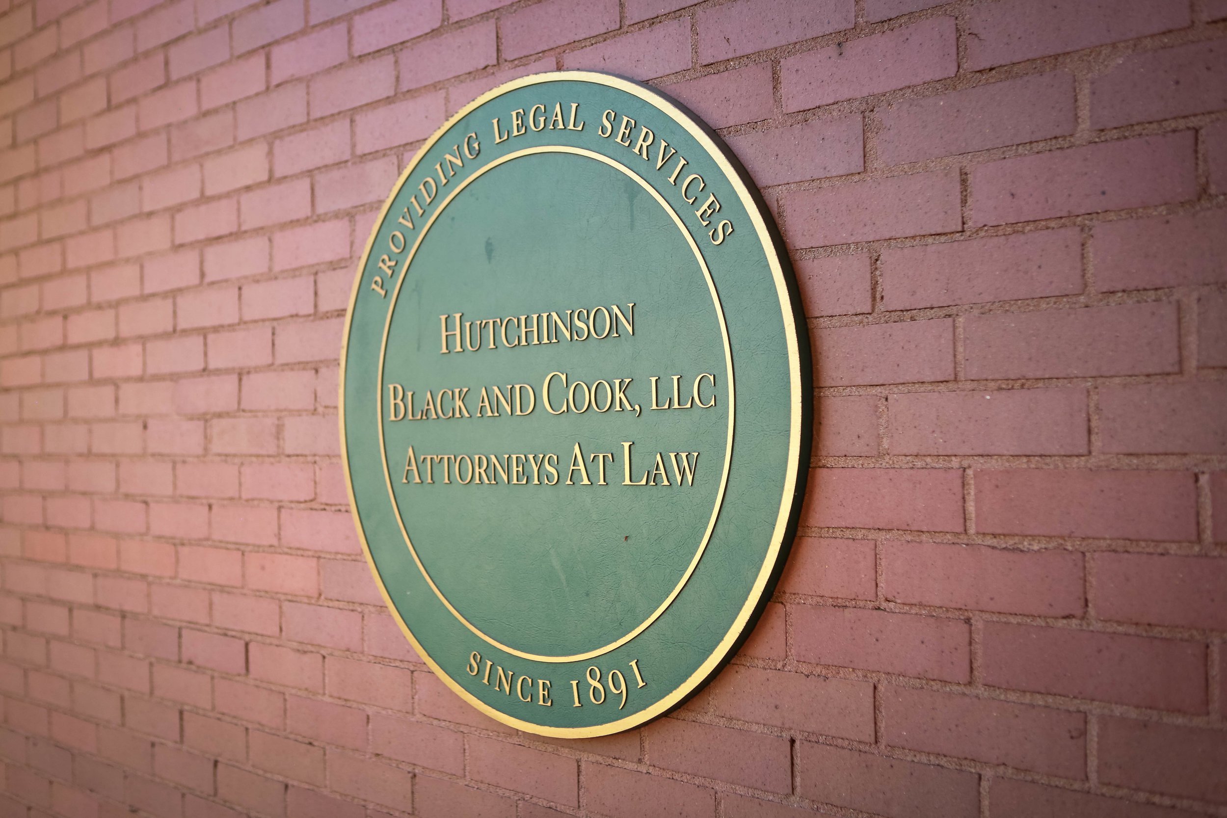 A close-up shot of a green, circular sign on a brick wall. The sign reads: "PROVIDING LEGAL SERVICES," "HUTCHINSON BLACK AND COOK, LLC ATTORNEYS AT LAW," and "SINCE 1891."