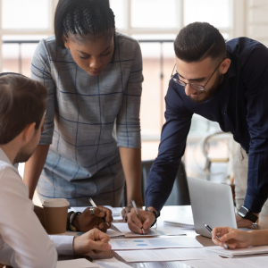 A diverse group of four professionals are gathered around a table with a laptop and documents, appearing to be collaborating on a project or reviewing data.