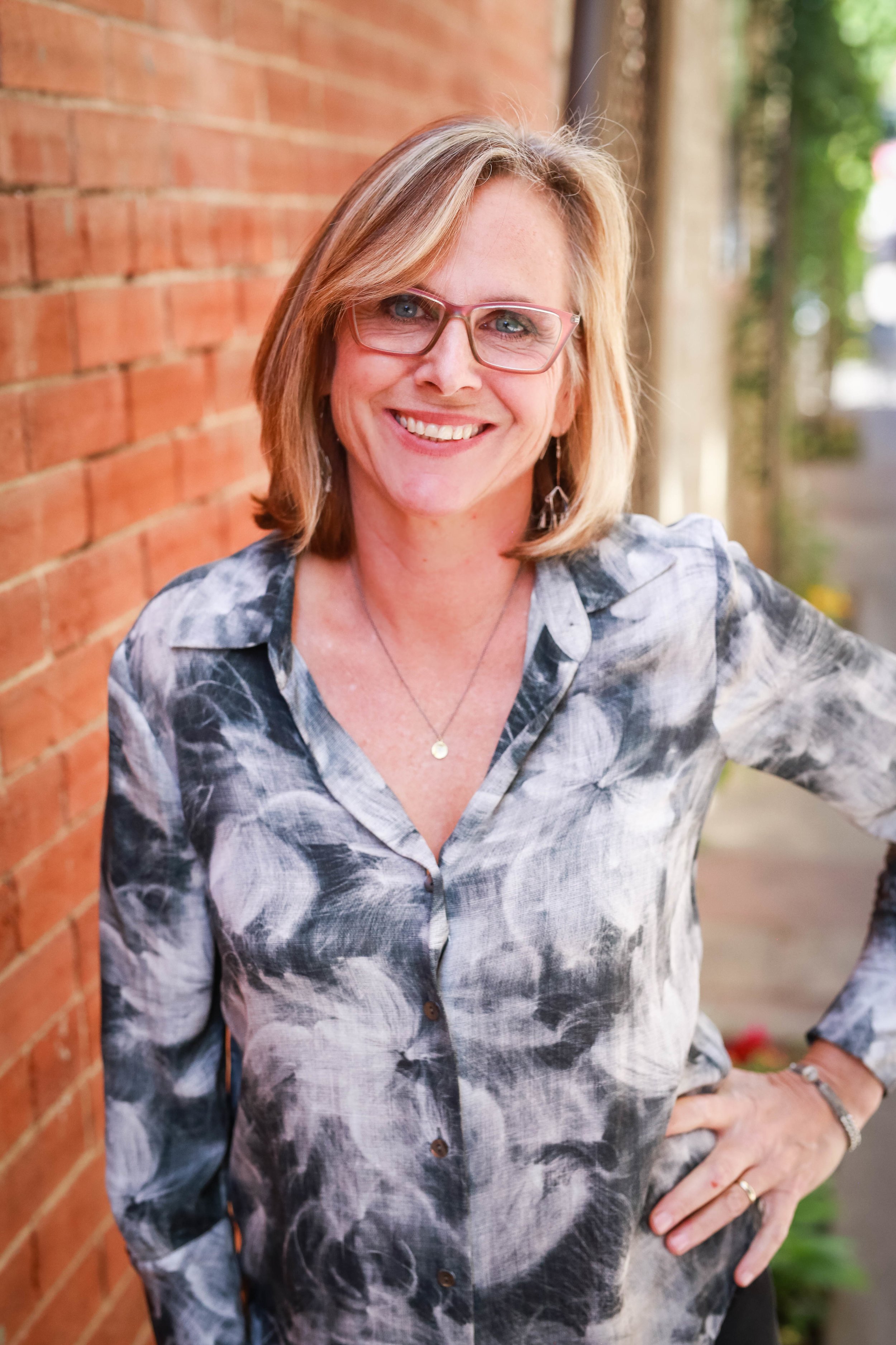 Professional headshot of blonde woman with glasses leaning against brick wall