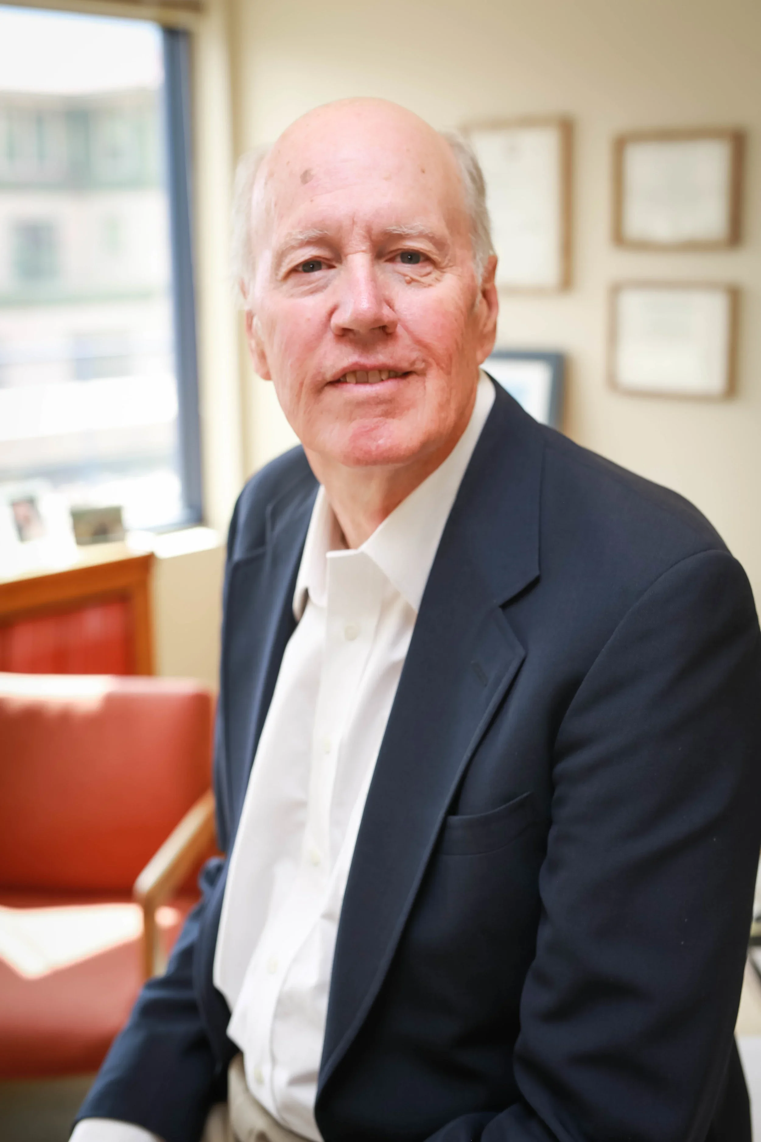 A professional headshot of a smiling senior man, wearing a dark suit jacket over a white shirt, seated in an office with a window and framed diplomas in the background.