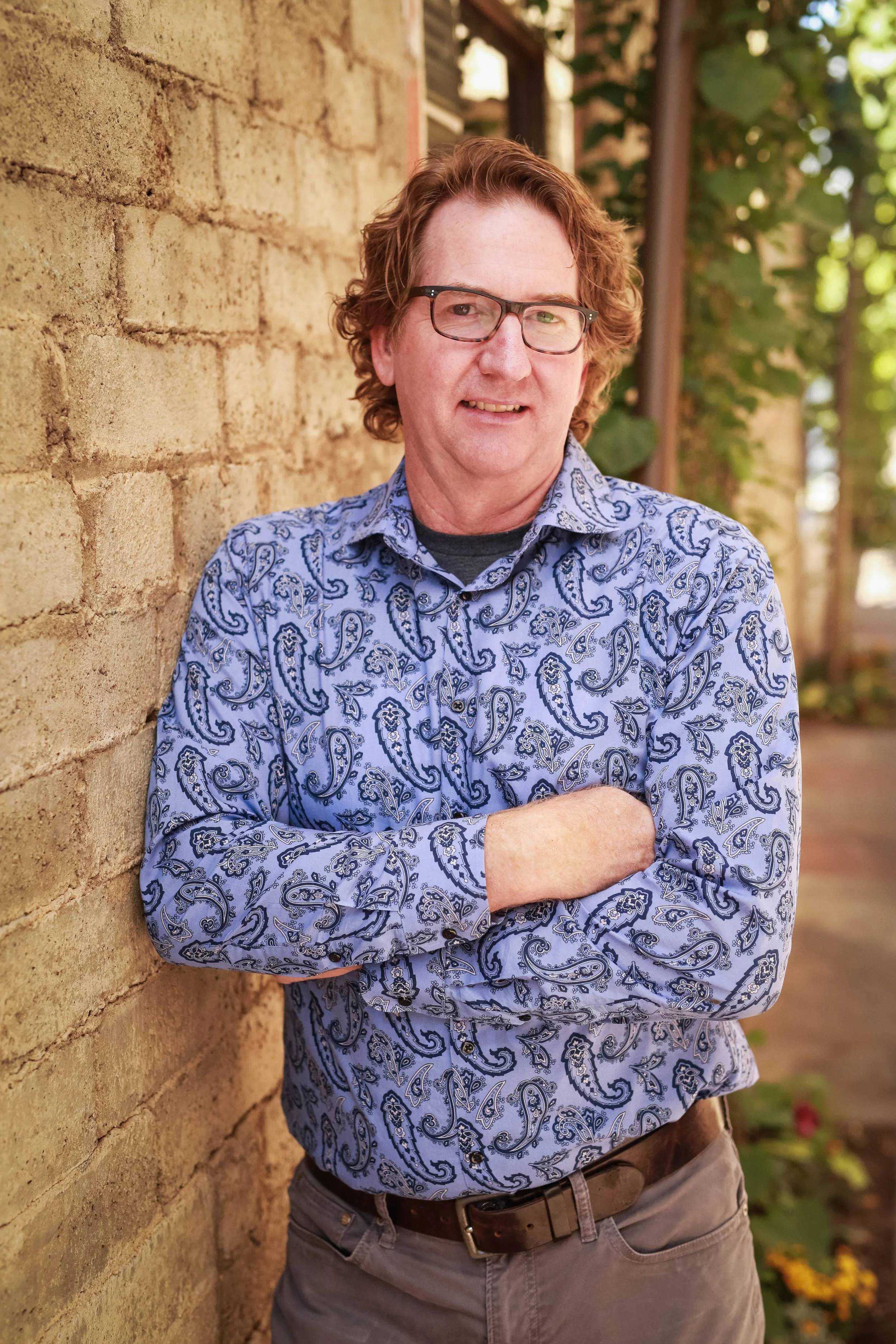 Professional headshot of man wearing paisley shirt, leaning against brick wall