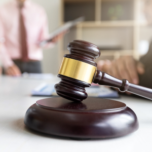 A judge's gavel and a scale of justice are on a white table, with blurred figures of people in business attire in the background, suggesting a legal meeting or court setting.
