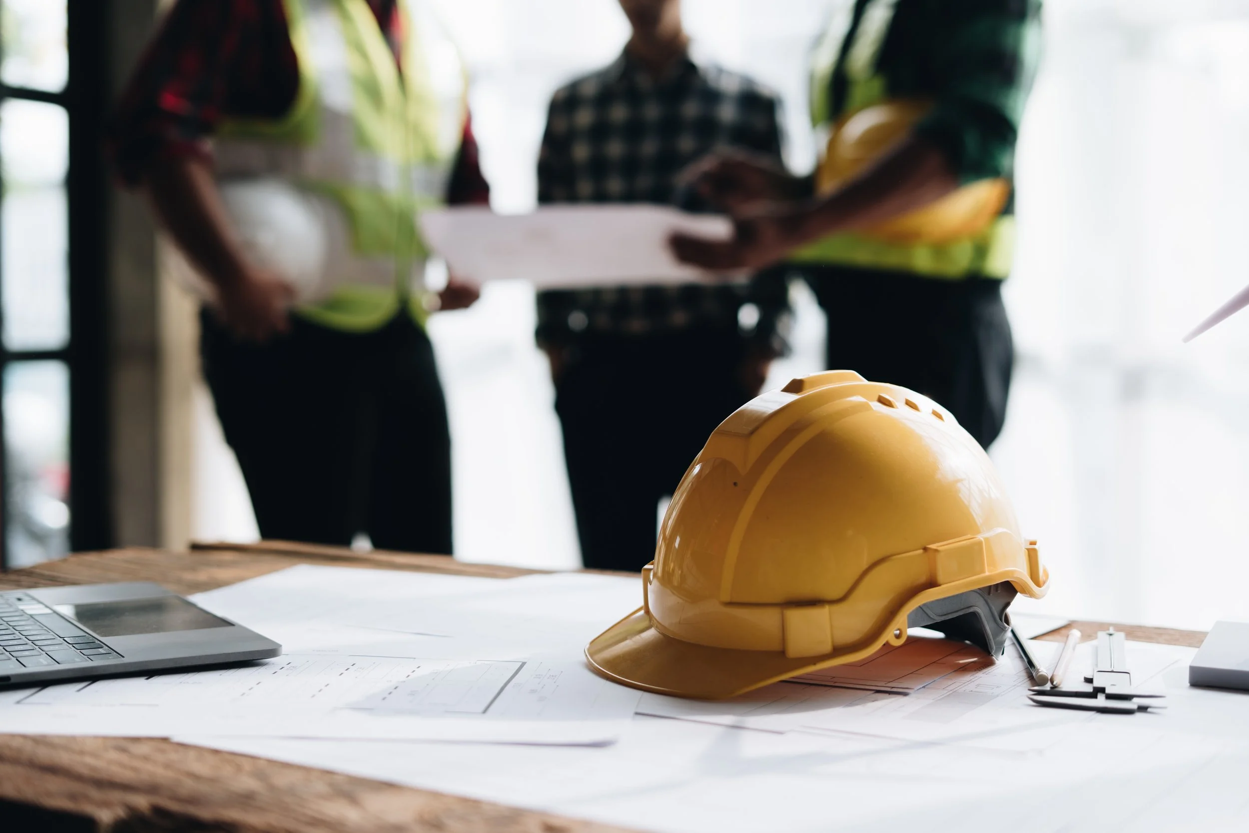 A yellow hard hat rests on architectural blueprints and a laptop, with three blurred figures in construction vests and helmets discussing plans in the background.