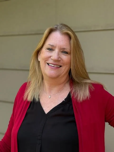 A smiling woman with long blonde hair, wearing a black shirt and a red cardigan, stands outdoors in front of a light-colored siding background.