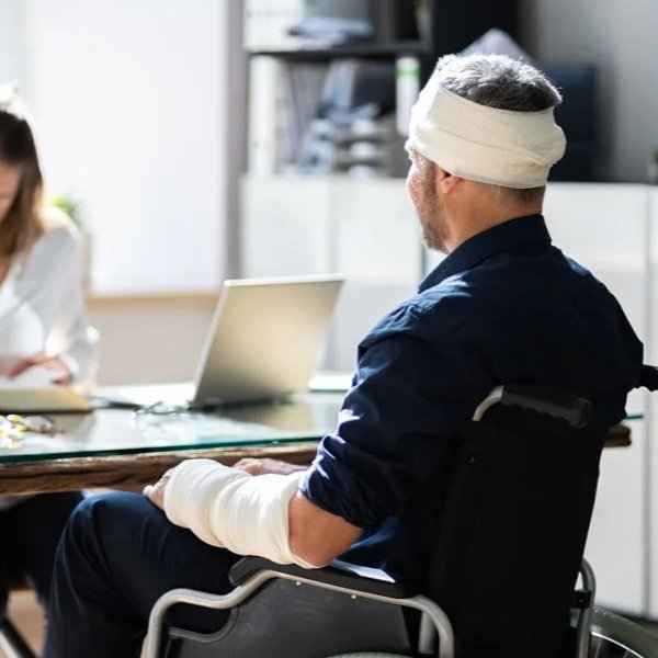 A man with a bandaged head and arm, in a wheelchair, is shown from behind facing a woman who is seated at a desk with a laptop and legal documents.