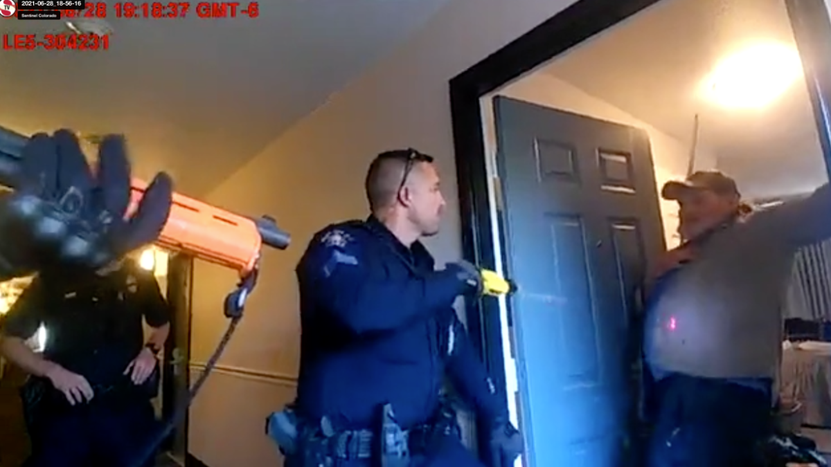 A blurry still image from a bodycam shows two police officers, one holding a less-lethal shotgun, at the threshold of a doorway where a man is partially visible. Time and date information are overlaid in the upper left corner.