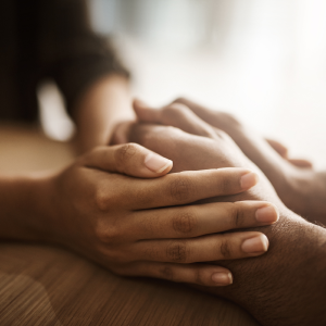 A close-up of two people's hands clasped on a wooden table, suggesting comfort, support, or empathy.