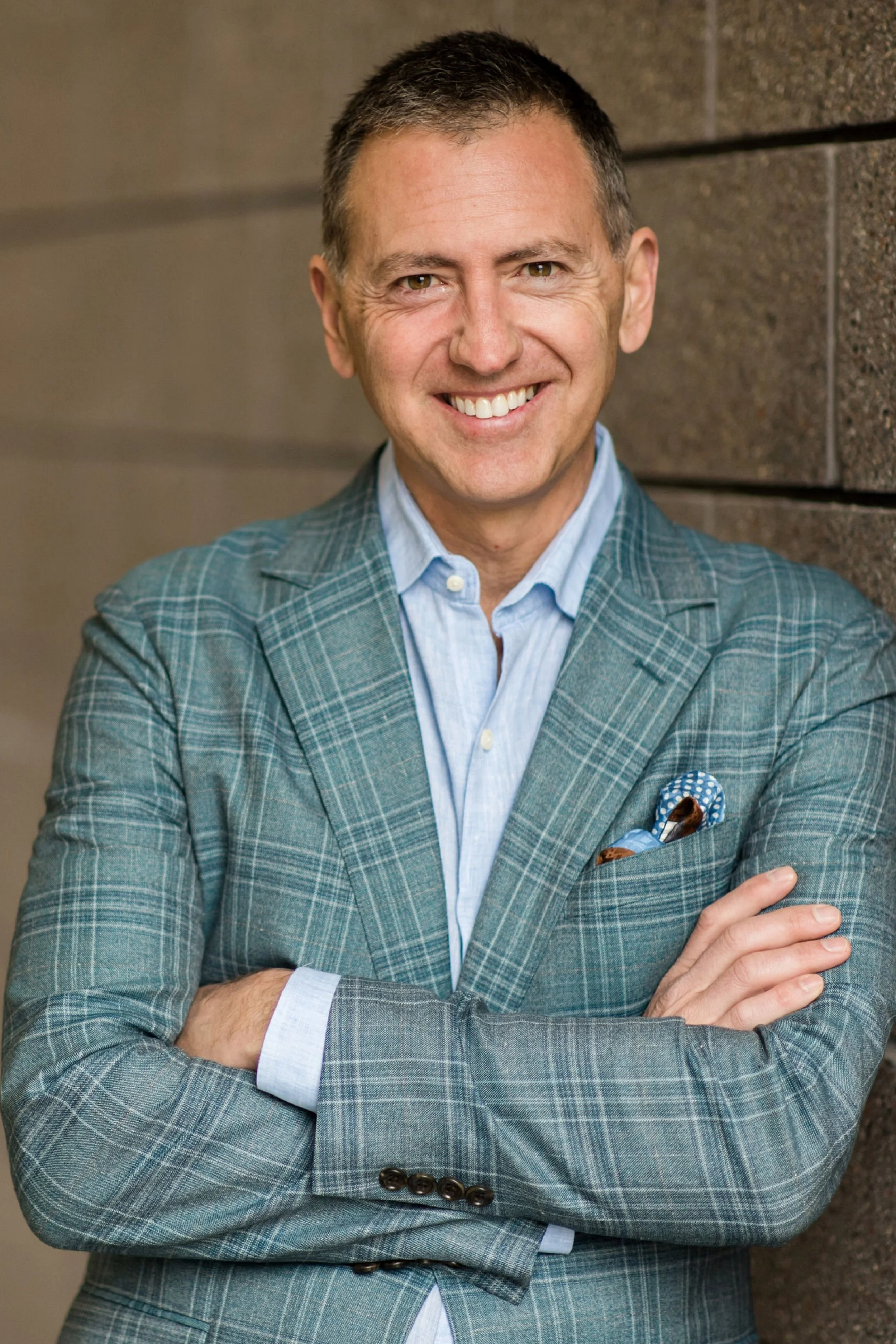 Profesional headshot of man wearing grey plaid suit leaning against wall