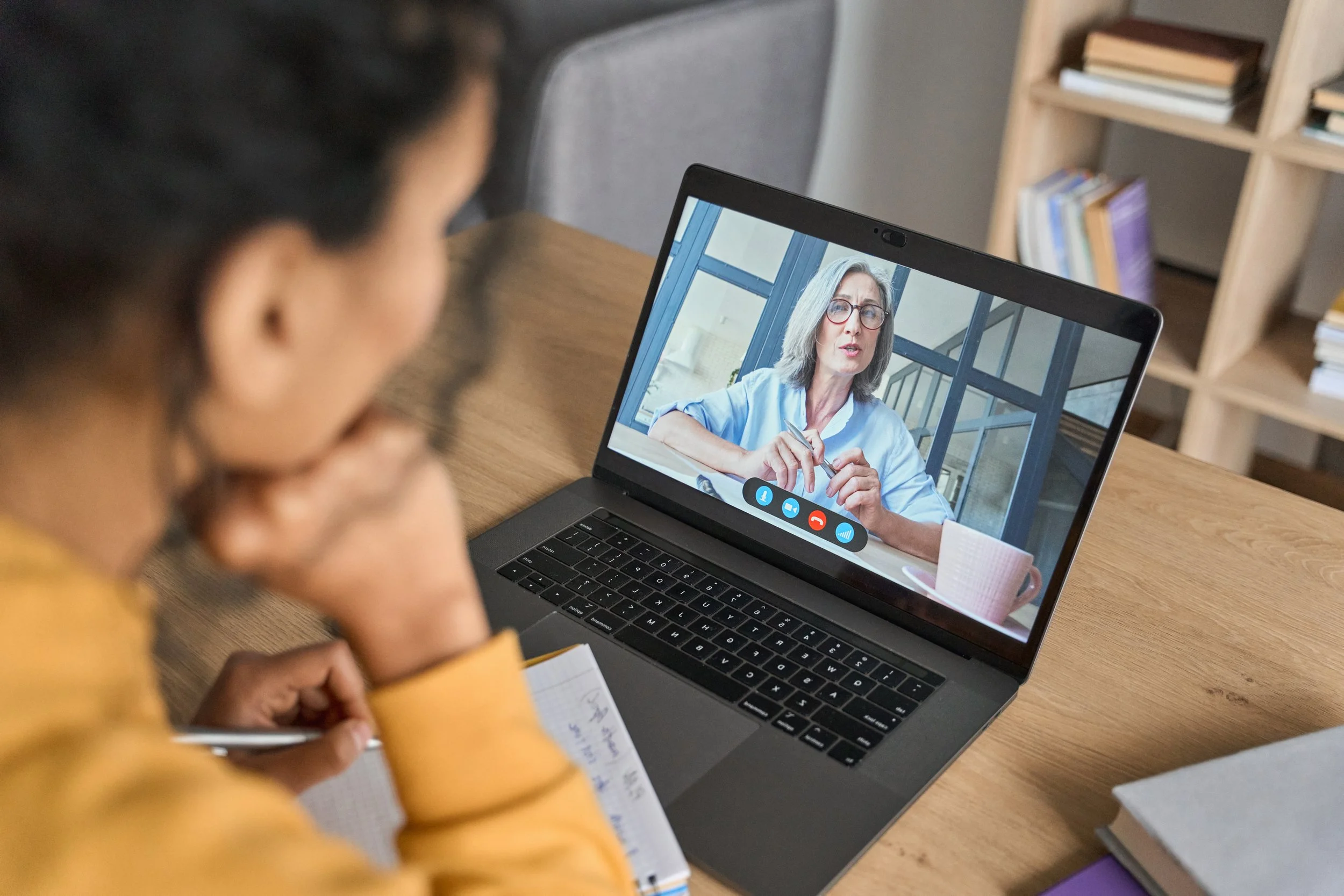 A person sitting at a wooden desk on a video call with a woman on a laptop screen. The woman on the screen has gray hair, glasses, and is speaking. The person at the desk is writing notes in a notebook and holding a pen.