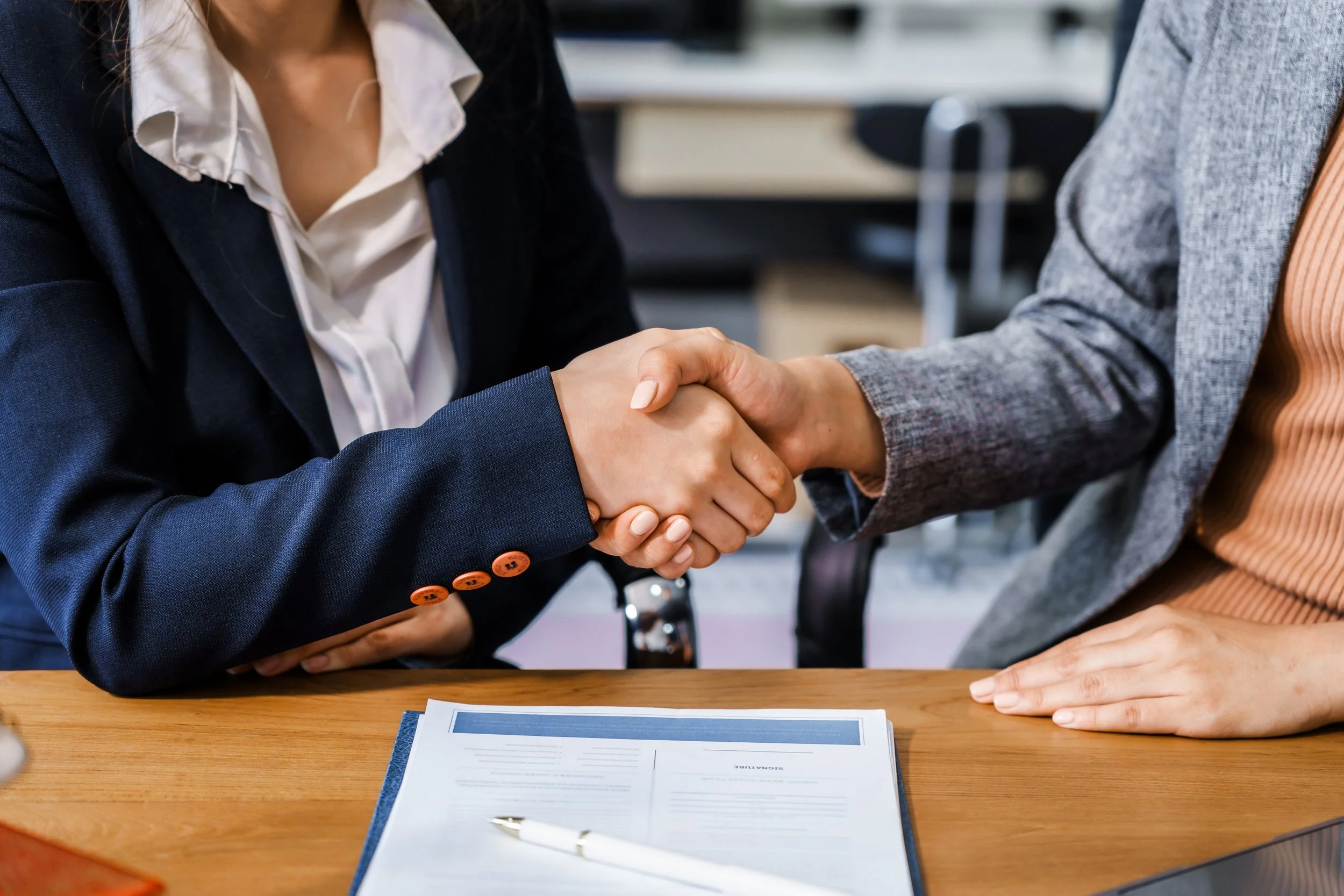Two women in business attire shake hands over a wooden table with documents and a pen.