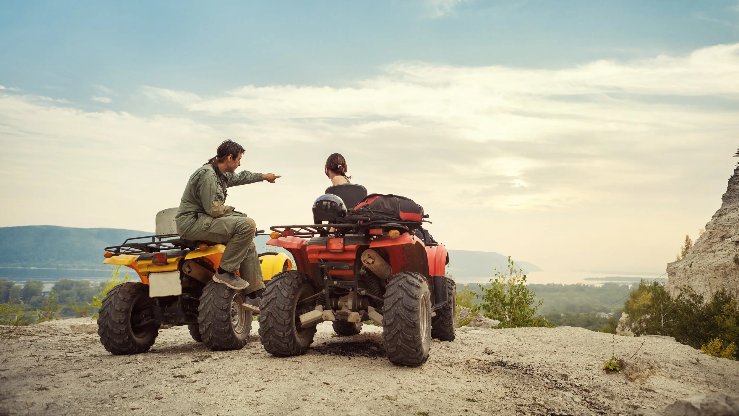 A man and woman sitting on ATVs on a rocky hill with a scenic view of a river and mountains in the background, during a cloudy day.