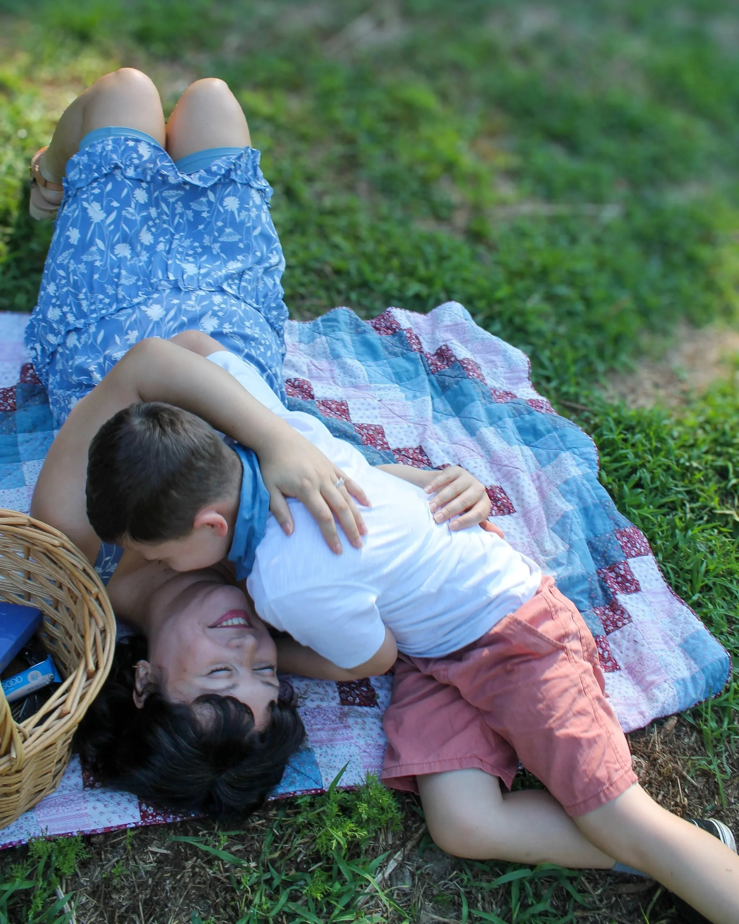 A woman and two children lying on a quilt on the grass outdoors, with one child resting on the woman's back and another child lying on their stomach in front of her, all appearing happy.