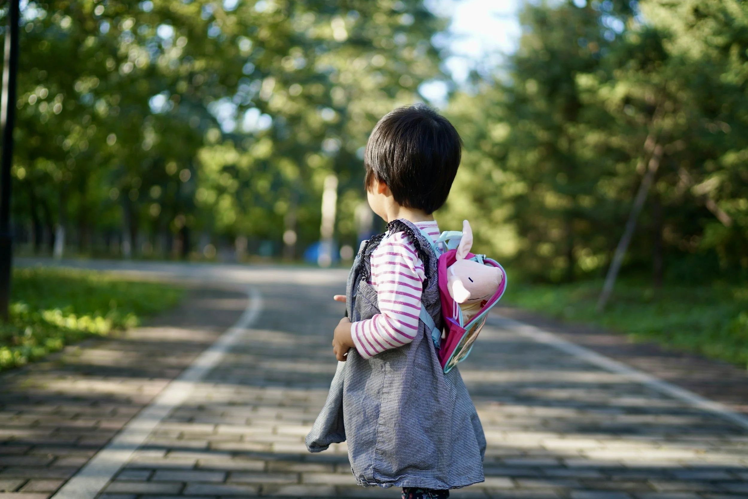 A young girl with short black hair wearing a pink and white striped shirt and a gray dress is walking outdoors with a colorful backpack featuring a plush doll in a pink section. She is on a paved path lined with green trees, looking to the left, in a park or wooded area during daytime.