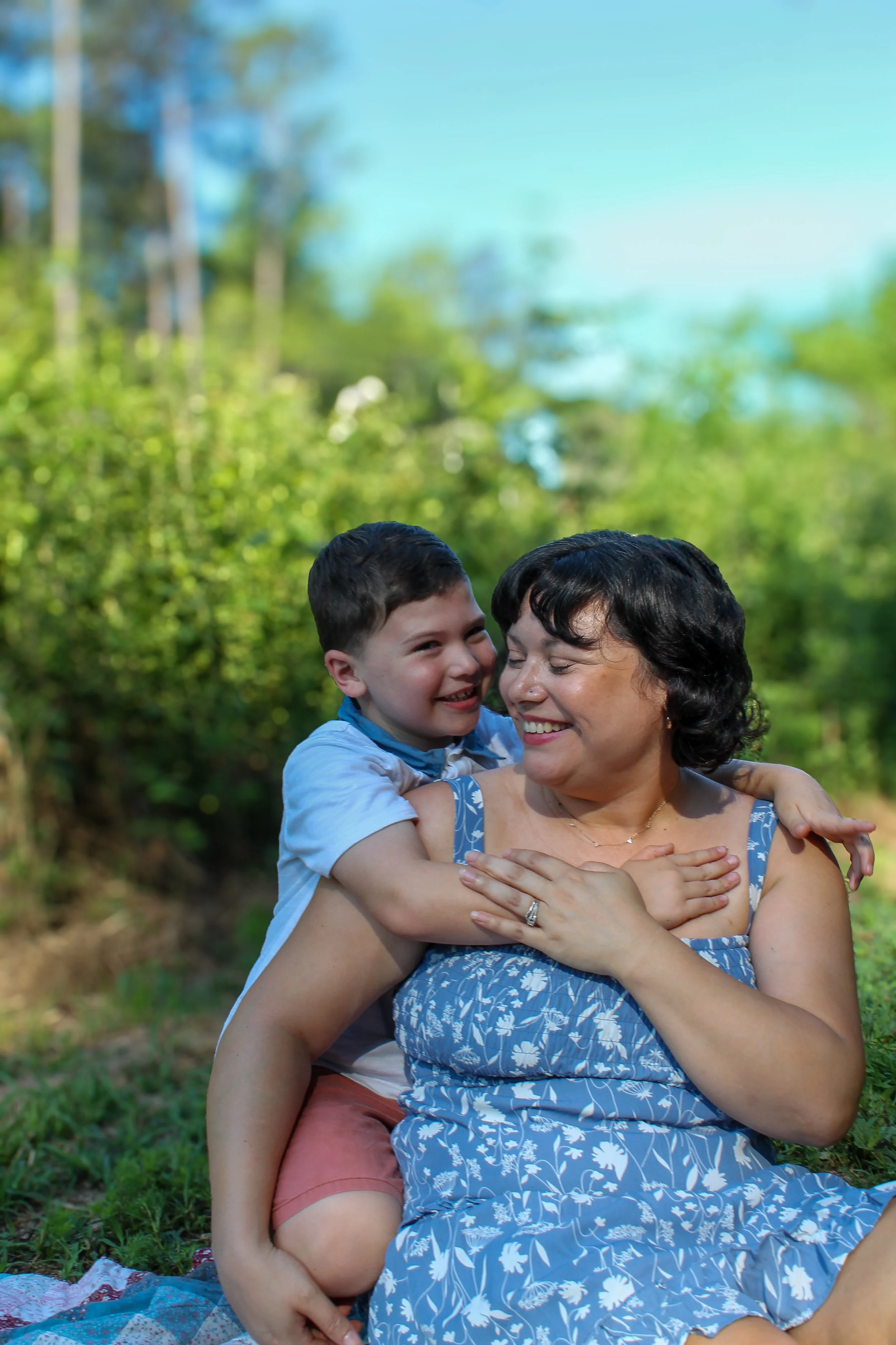 A woman with dark curly hair smiling and hugging a young boy with short dark hair, both outdoors with a blurred green forest background.