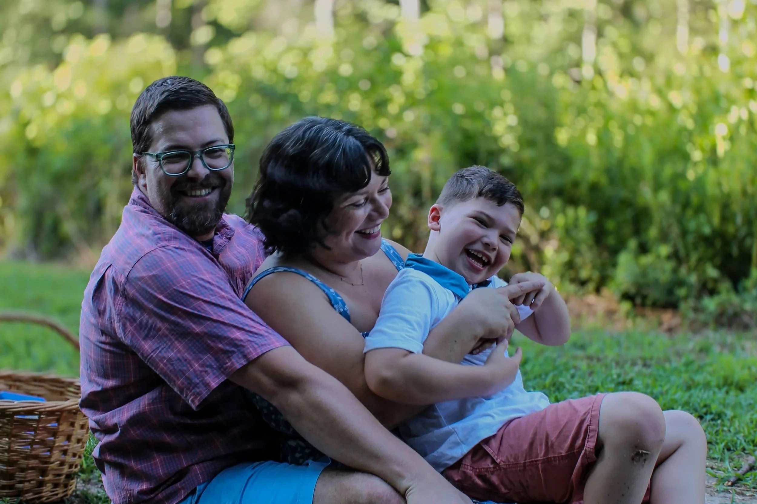 Three people, a man, a woman, and a boy, sitting outside on the grass, laughing and smiling, with trees in the background. A wicker basket is next to them.