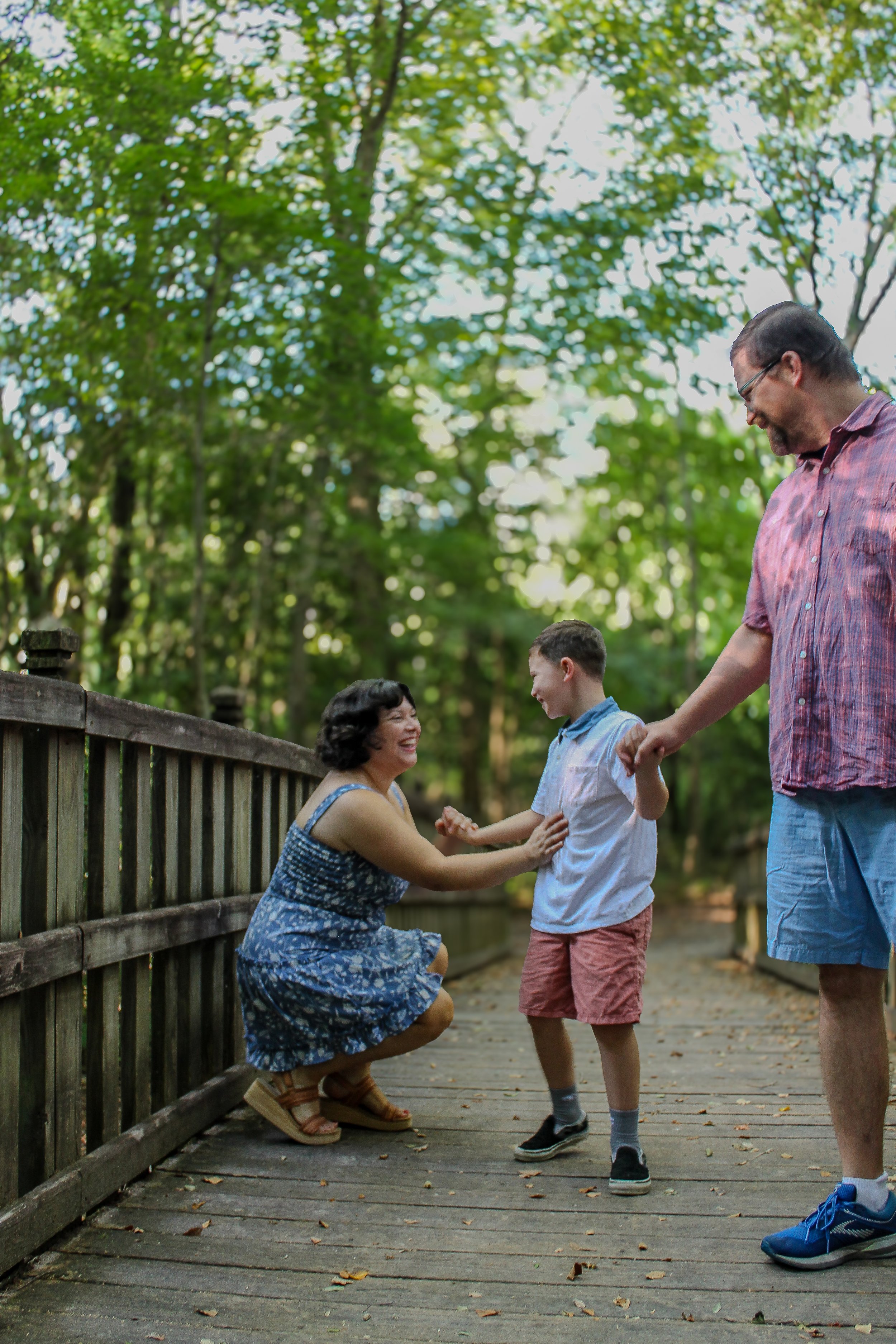 A woman kneels on a wooden bridge, smiling and holding hands with a young boy who is standing in front of her. A man, smiling, stands nearby holding the boy's hand. They are outside amidst green trees with sunlight filtering through the leaves.