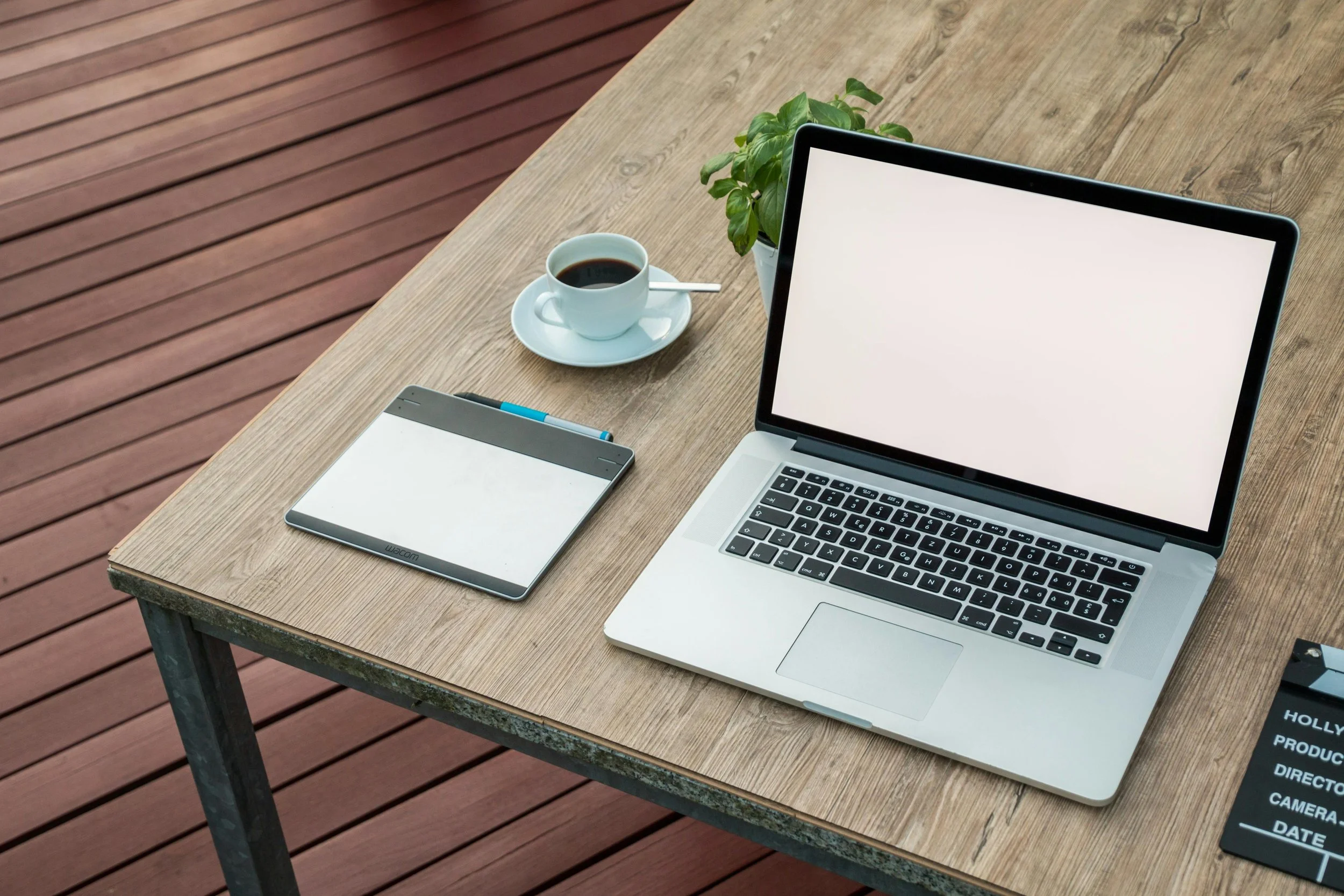 A wooden outdoor table with a laptop, a tablet, a cup of black coffee on a saucer with a spoon, a green potted plant, and a smartphone on a wooden deck.