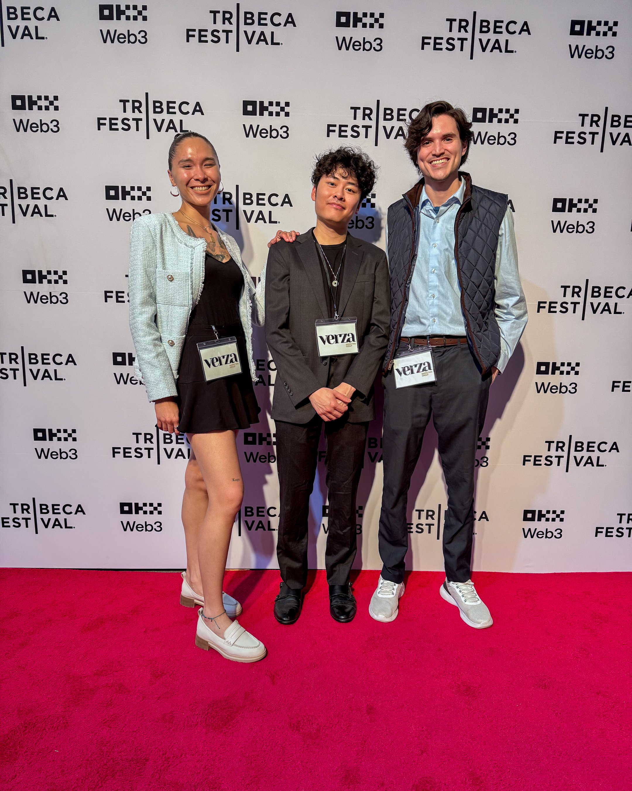 Three people standing on a red carpet at the Tribeca Festival Web3 event, smiling for the camera. A woman on the left wears a light blazer, black dress, and white shoes. A man in the middle wears a black blazer, black shirt, and black shoes. A man on the right wears a light blue shirt, dark vest, and white sneakers. The backdrop features the Tribeca Festival Web3 logo repeatedly.
