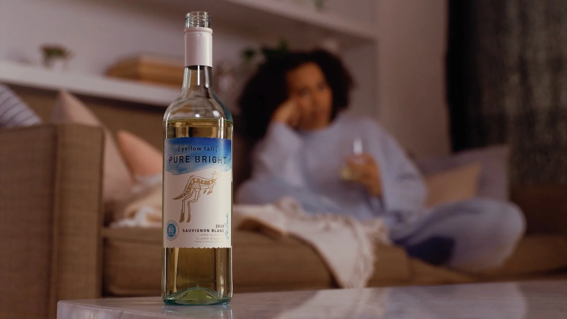 A bottle of Yellow Tail Pure Bright Sauvignon Blanc white wine placed on a table in the foreground. In the background, a woman with curly hair and a gray shirt is sitting on a bed, looking relaxed and holding a glass of wine.