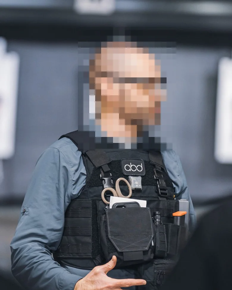 A person wearing a tactical vest with various tools and items, including scissors, a phone, and a flashlight, with a blurred background.