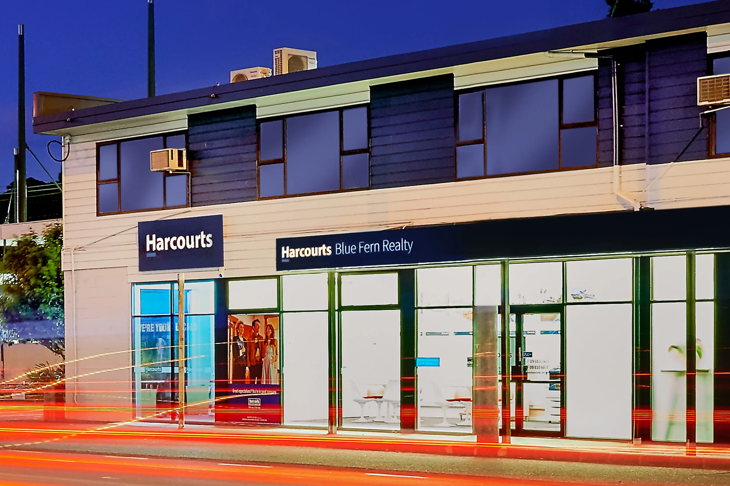 Exterior view of a real estate agency named Harcourts Blue Fern Realty at dusk, with light trails from passing cars in front.