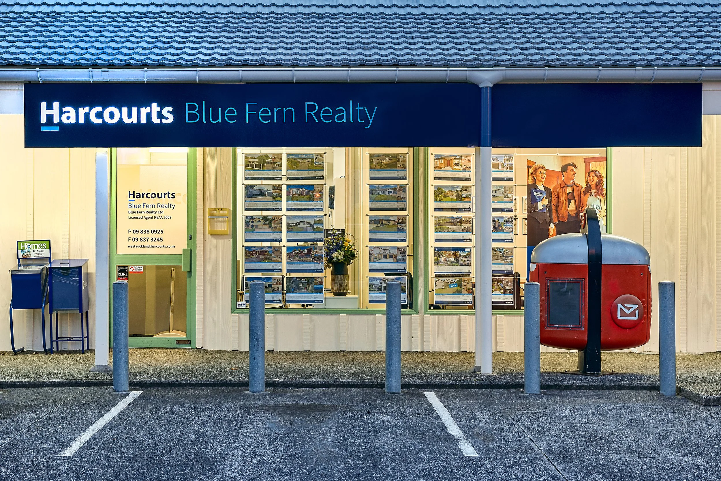 Real estate office with Harcourts Blue Fern Realty sign, display of property photos in window, and a red mailbox outside.