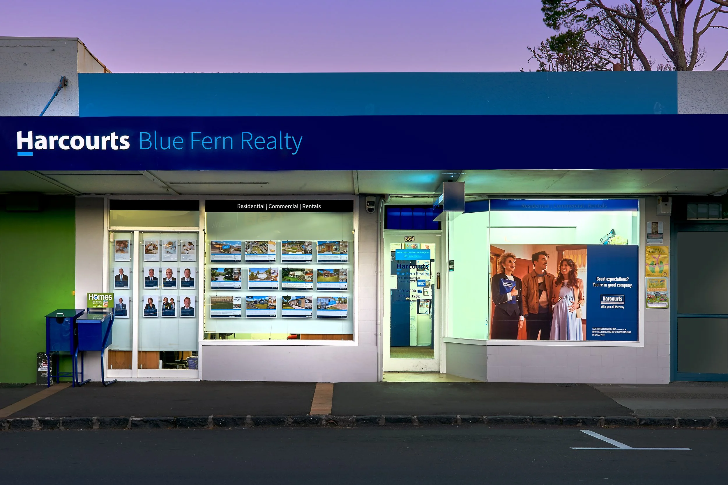 Harcourts Blue Fern Realty real estate office with display windows showing property listings, a large poster of three people in a home, and a blue sign with the company name on the building.