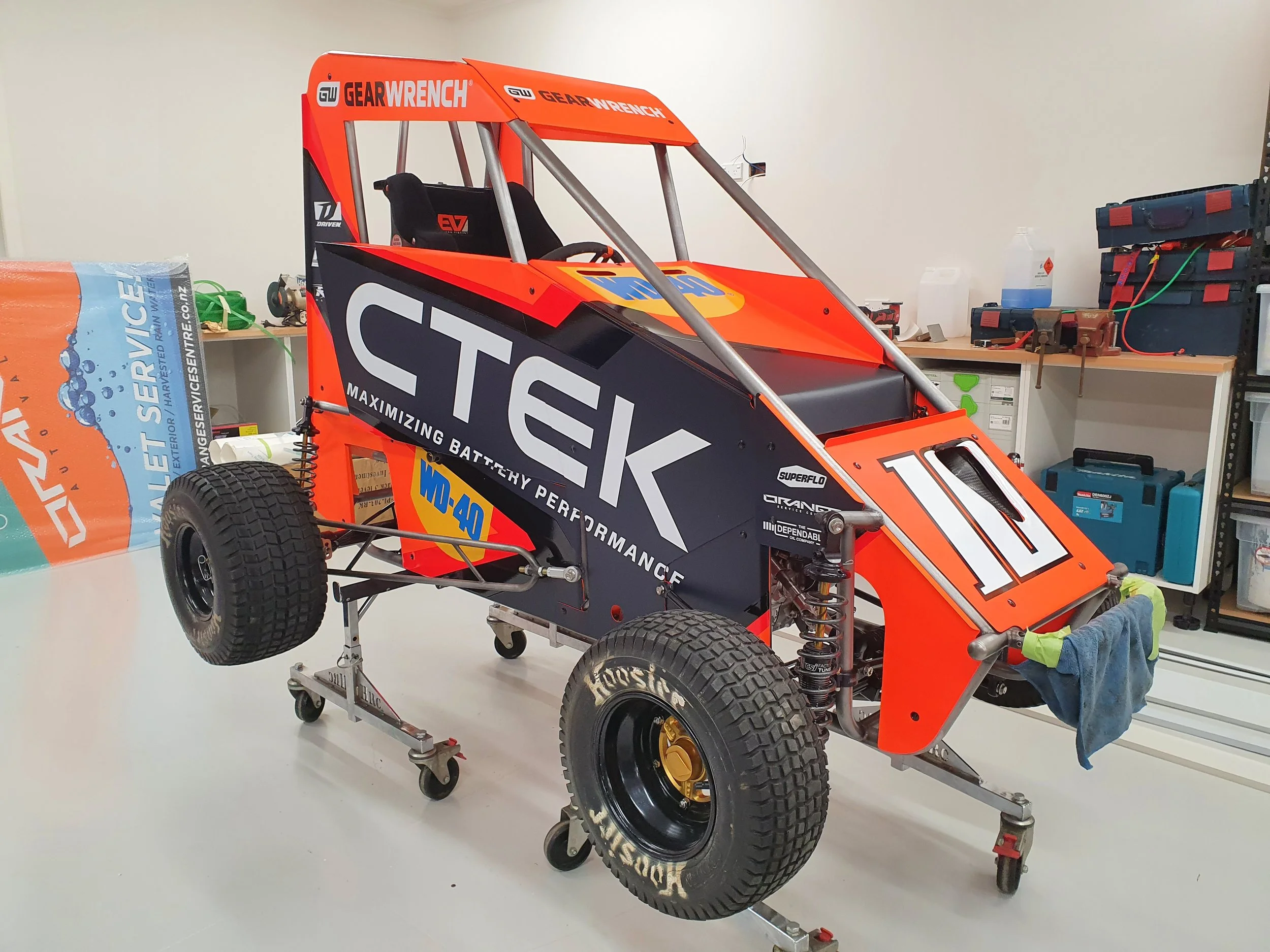 A race car chassis with sponsor decals, mounted on a wheeled stand, in a workshop with tools and storage shelves in the background.