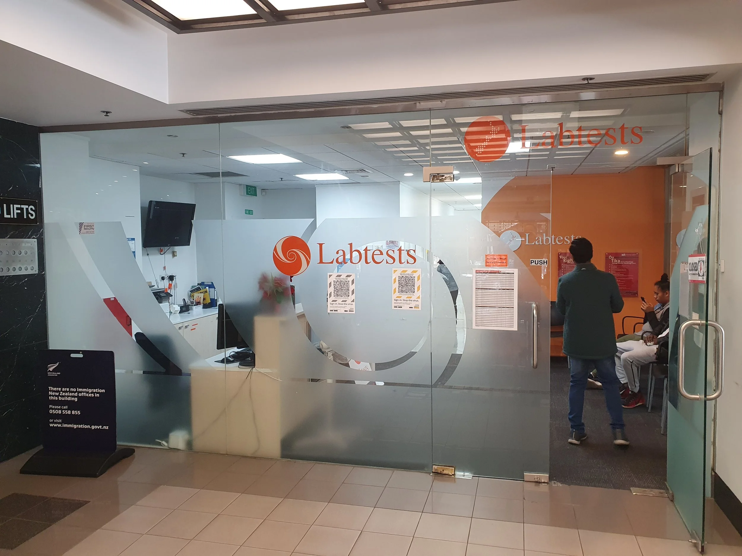 A glass entrance to a lab testing clinic named 'Labtests' with chairs and people inside, some waiting and one standing near the door. The entrance has the lab's logo and QR codes on the glass.