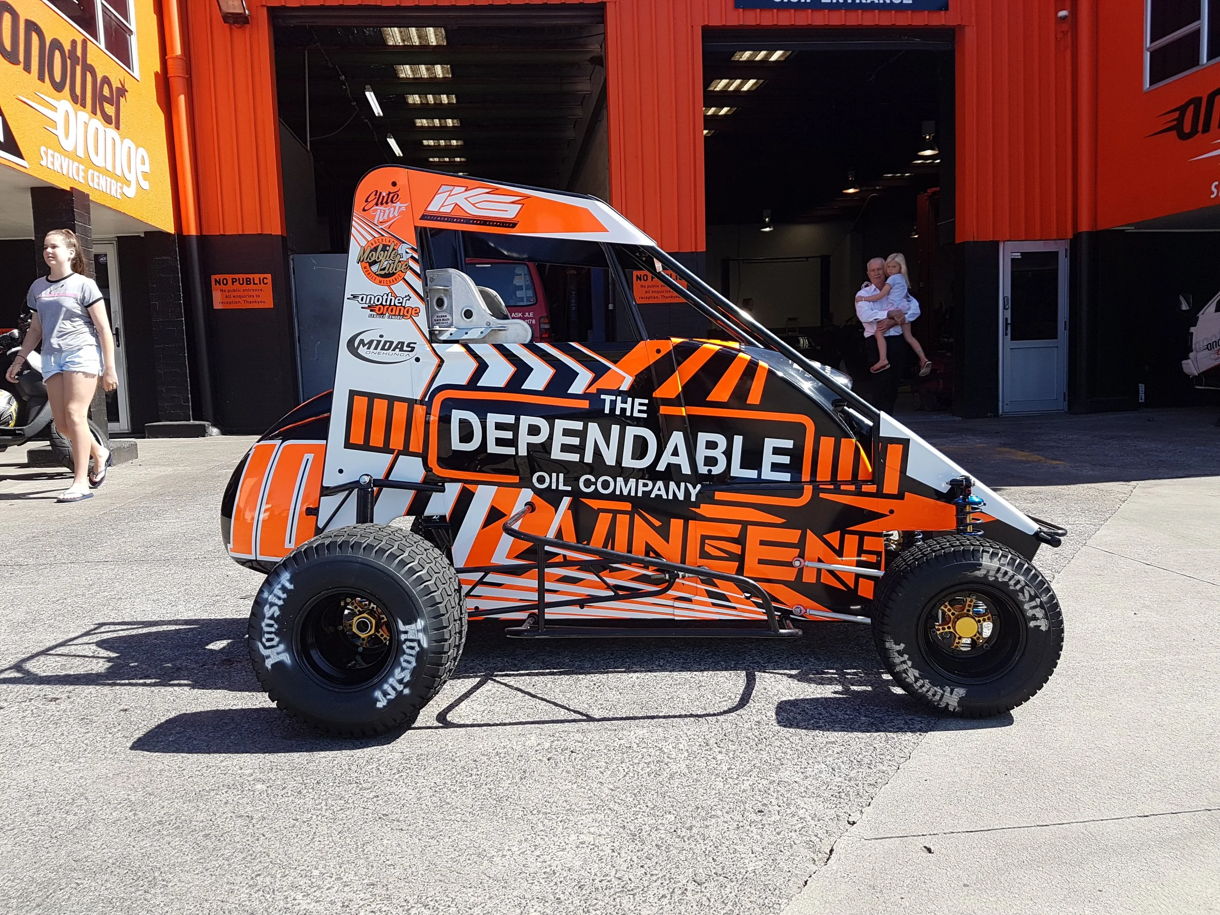 A race car with orange, black, and white graphics sponsored by The Dependable Oil Company parked outside a building with orange and black exterior walls. Two women and a man with a child walk and stand near the car.