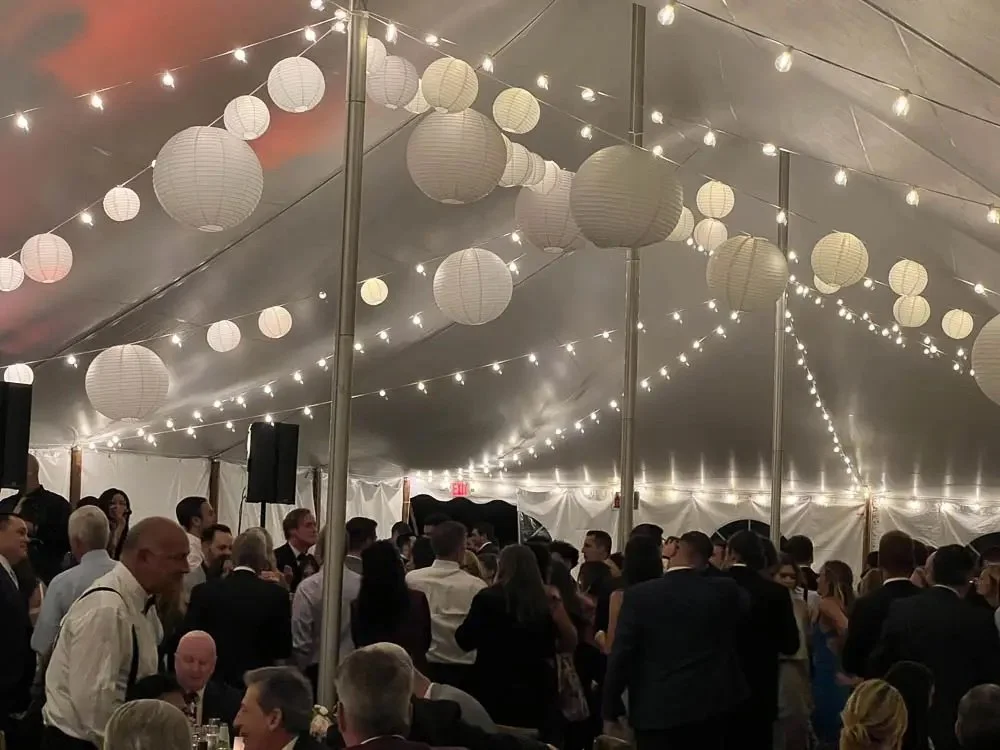 People dancing and socializing inside a decorated event tent with hanging paper lanterns and string lights.
