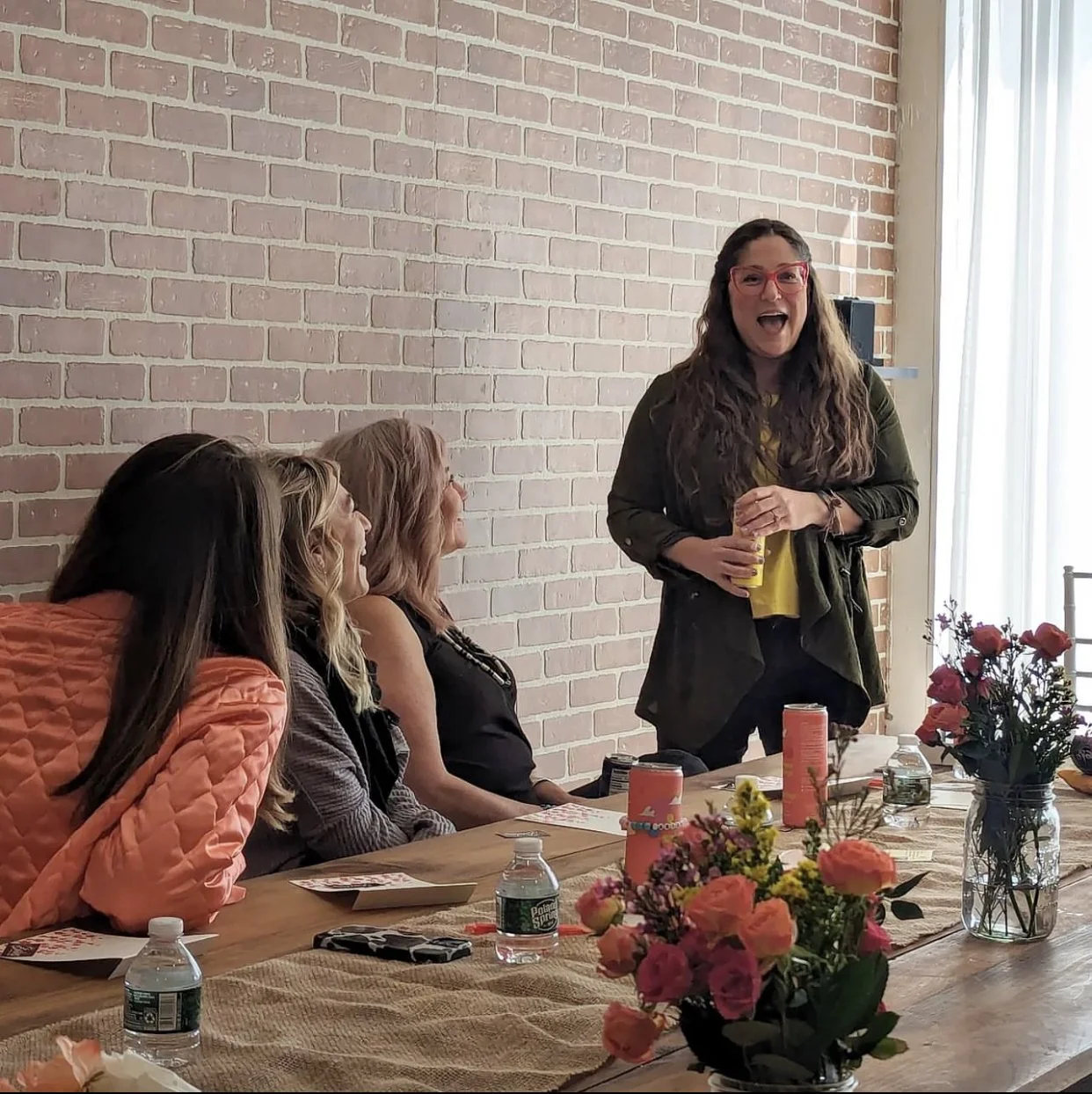 A woman with long curly hair, red glasses, and a yellow shirt standing and speaking to a group of three women seated at a table with flowers and water bottles, in a room with a brick wall and large windows.