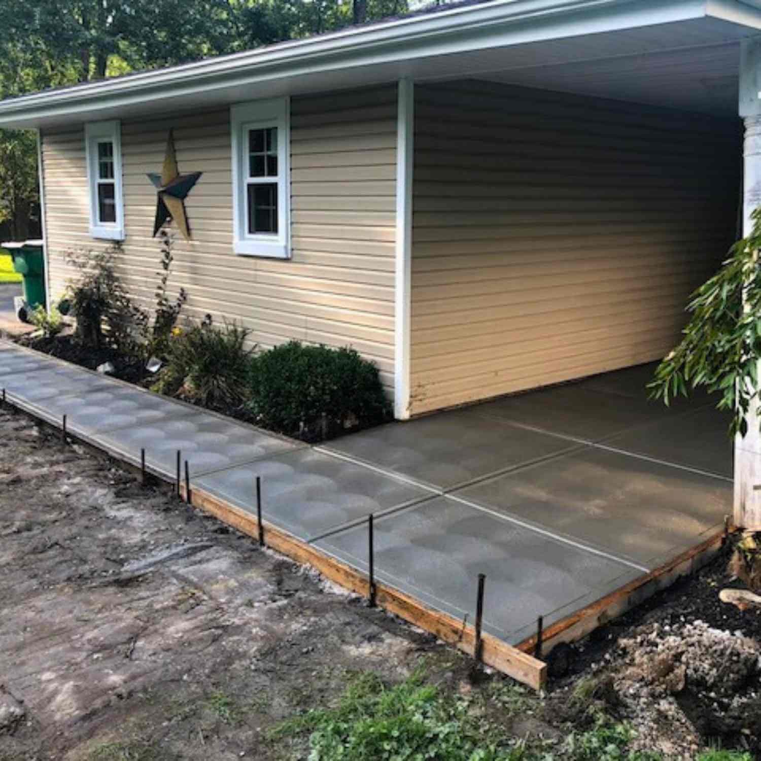 Newly poured concrete sidewalk and porch in front of a beige house with two windows, star decoration, and landscaping including bushes and a tree.
