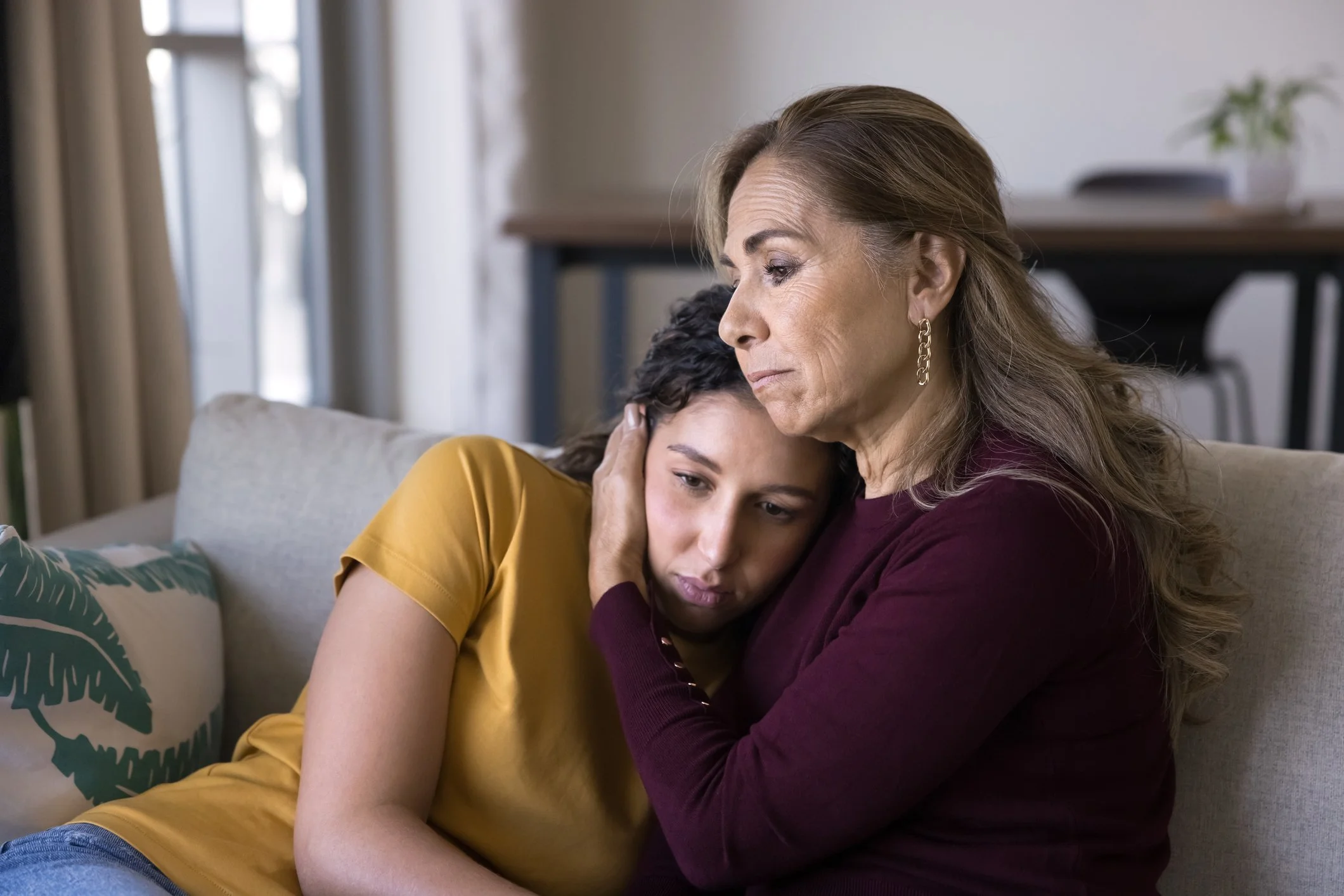 An older woman and a young girl sitting closely on a couch, comforting each other with the woman's arm around the girl and their heads touching, in a cozy living room with a plant and a table in the background.