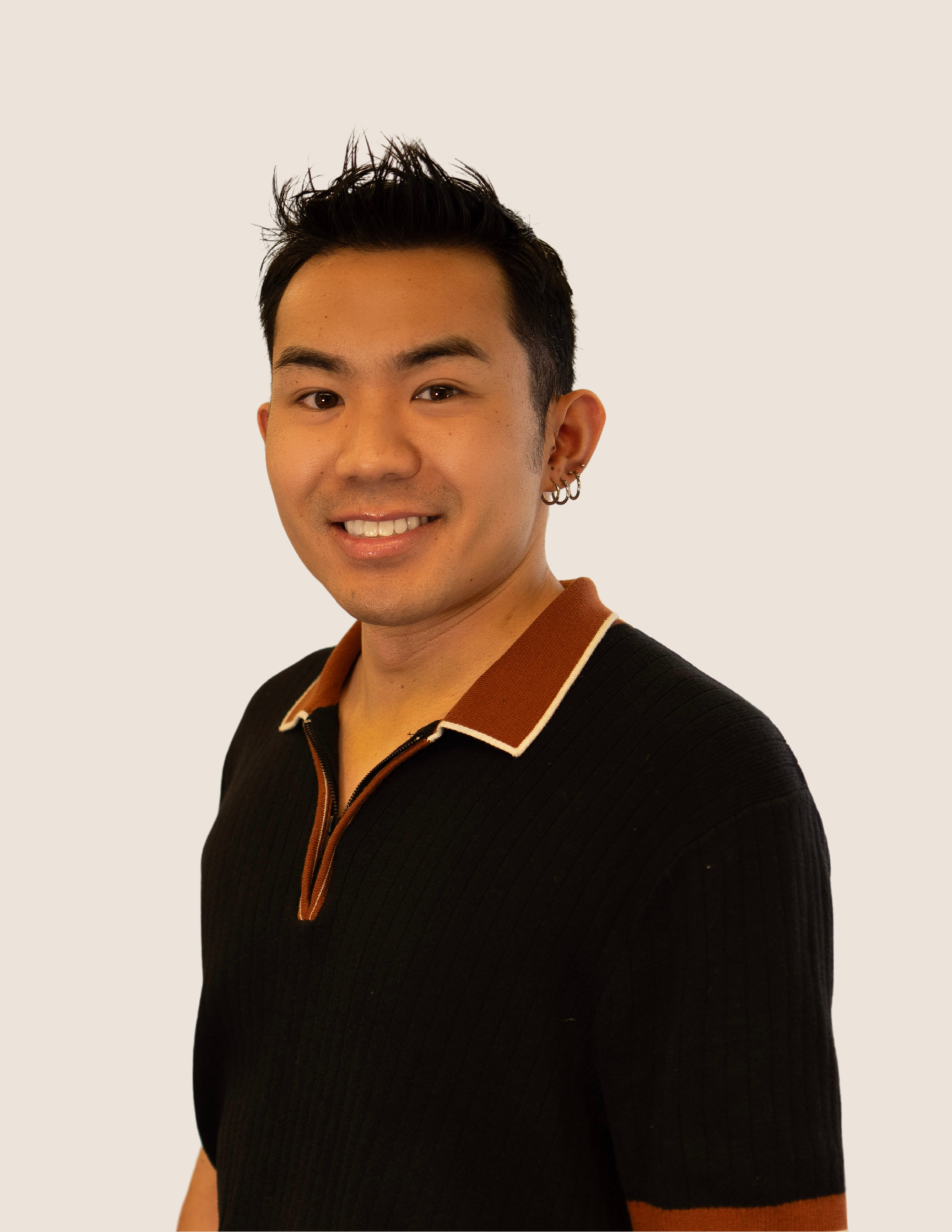 A young man with short black hair, wearing a black shirt with brown collar, smiling against a plain light background.