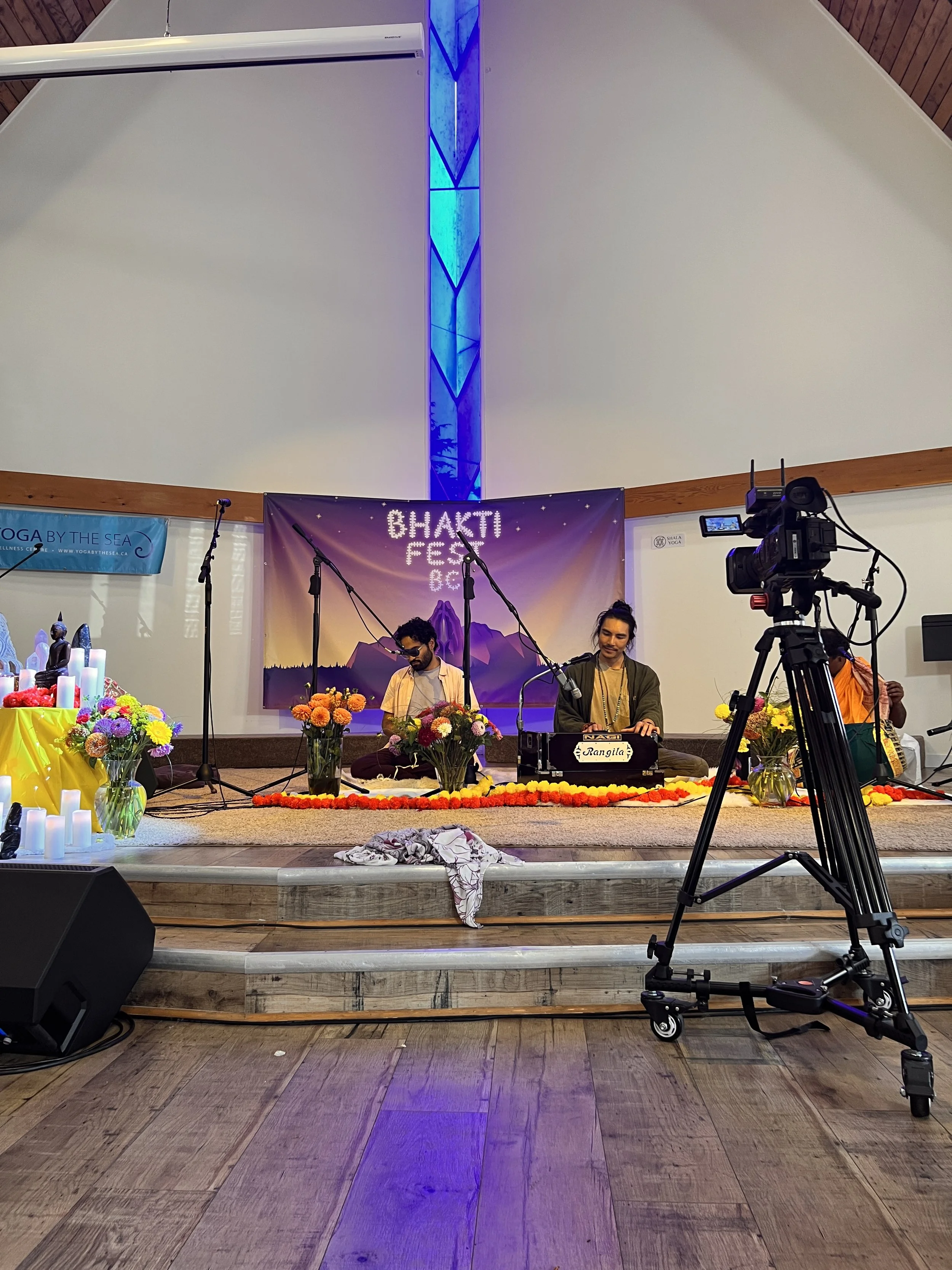 Two musicians performing on stage during Bhakti Fest, with one playing a harmonium and the other a keyboard, surrounded by floral arrangements and lit candles, with a camera recording the performance.