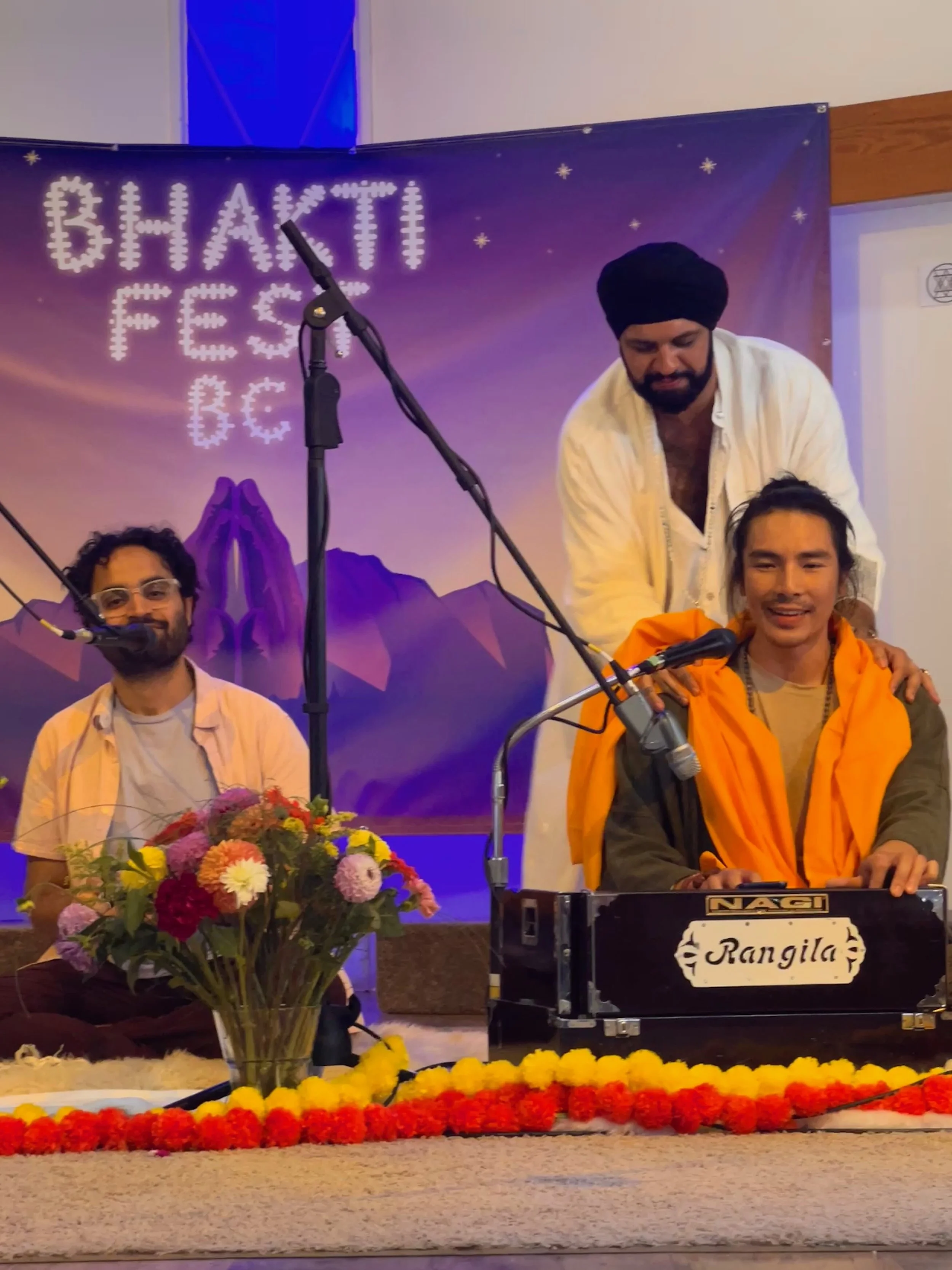 Three men are on stage at a cultural event with a banner in the background that reads 'Bhakti Fest'. One man in a white kurta and turban is standing behind a seated man wearing a bright orange shawl, who is playing a harmonium labeled 'Rangila'. The 