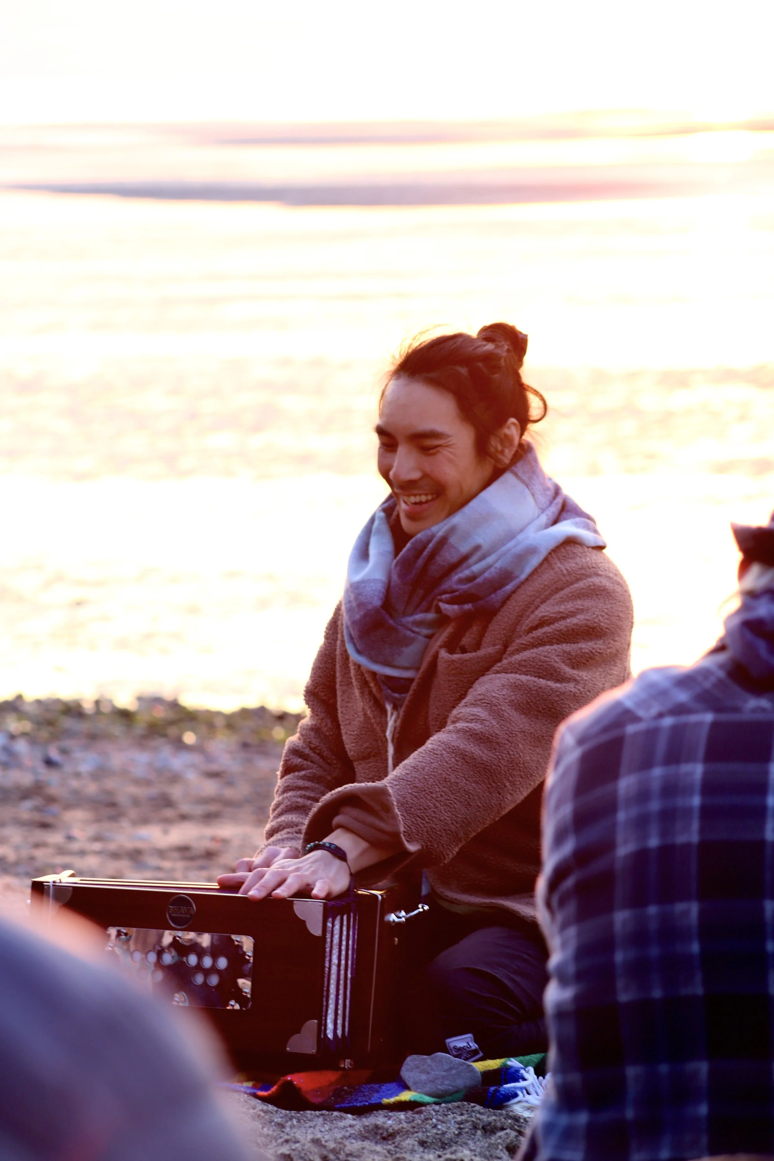 A man smiling and playing guitar on the beach at sunset, wearing a jacket and scarf.