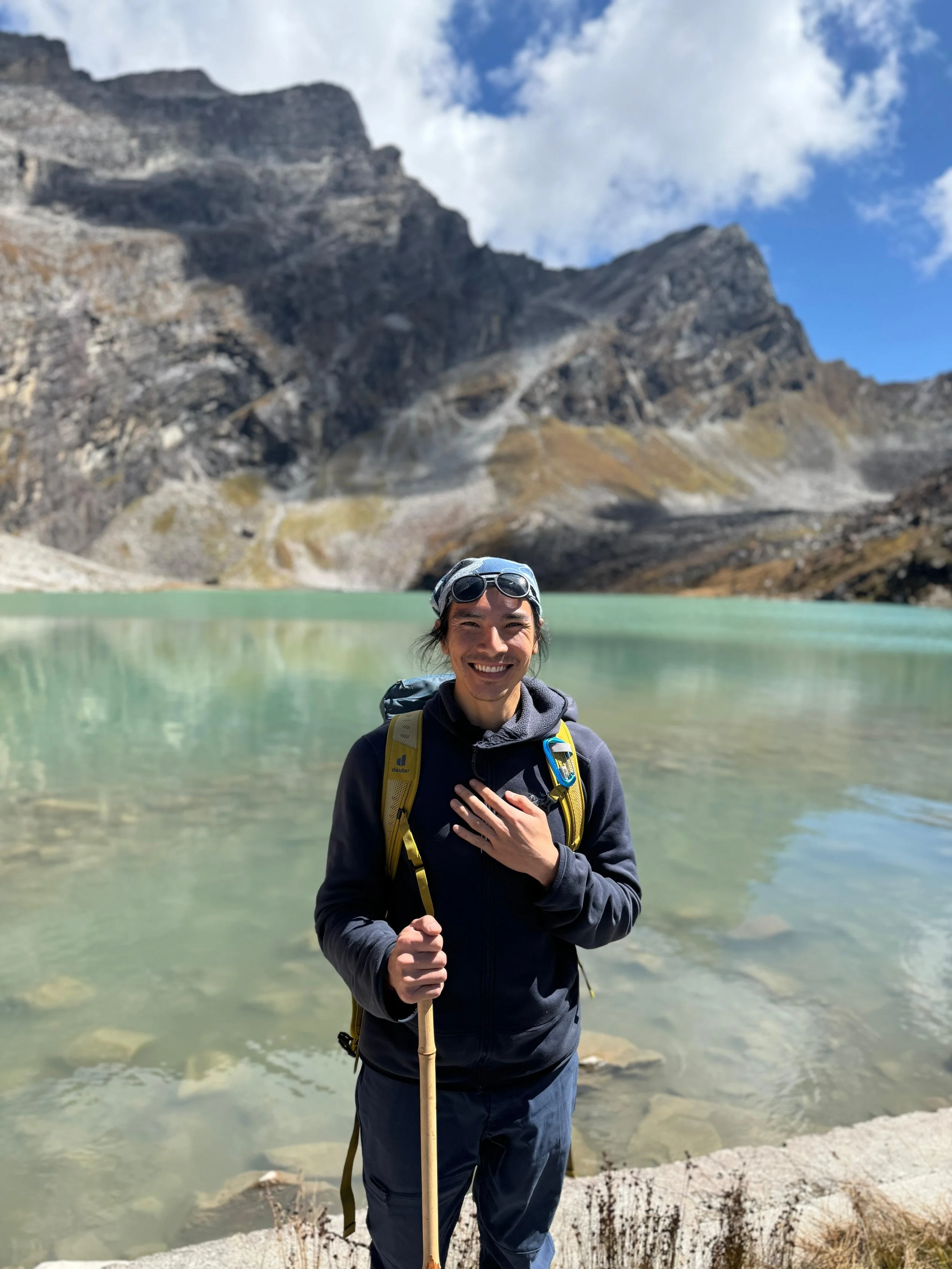 A man standing in front of a mountain lake, smiling and holding a walking stick, wearing a backpack, sunglasses, and outdoor gear with mountains in the background.