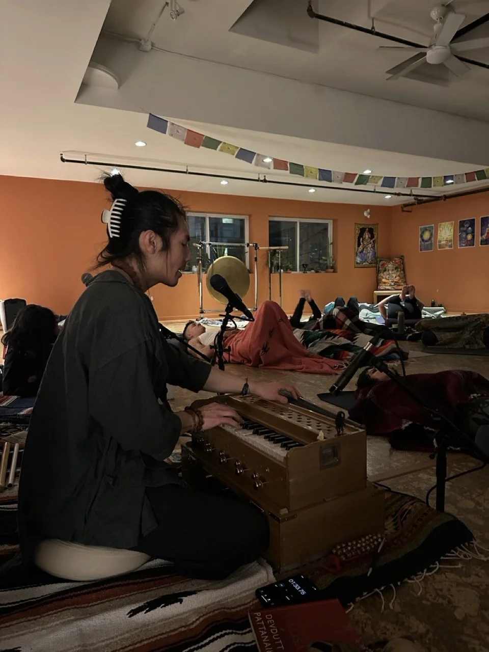 A person playing a harmonium in a yoga or meditation class with others lying on mats in a relaxing, dimly lit room.
